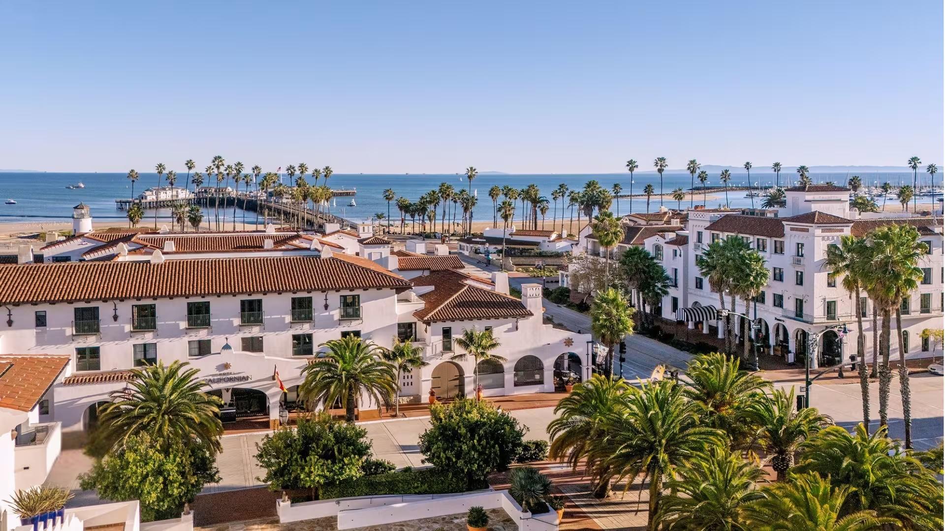 An aerial daytime view of a white Spanish-colonial hotel complex beside a palm-lined beach, a pier, and the ocean, with sailboats and a distant marina.