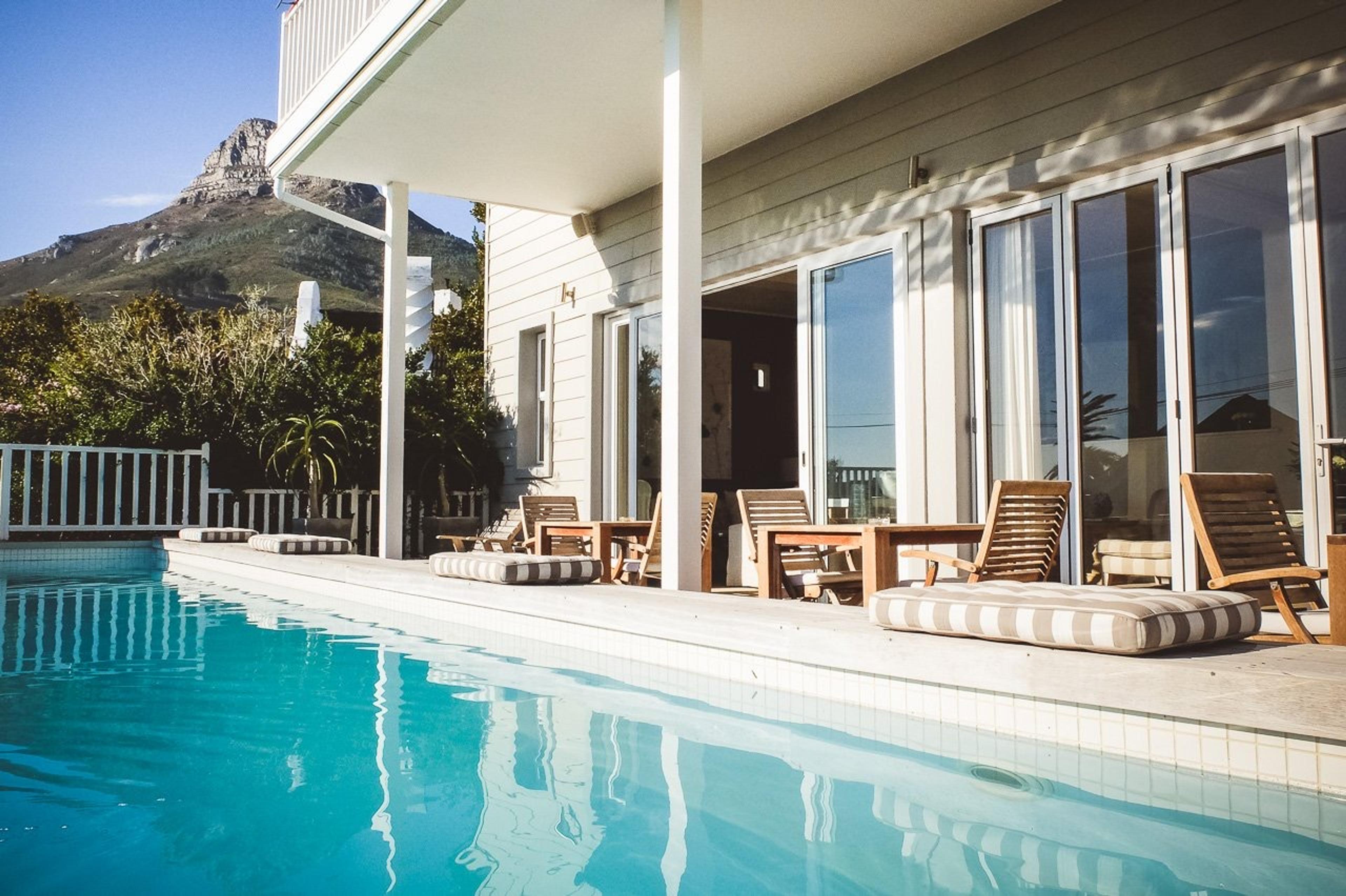 A white timber house seen from its pool, with a covered veranda, teak loungers with striped cushions, and a rocky mountain rising behind the trees.