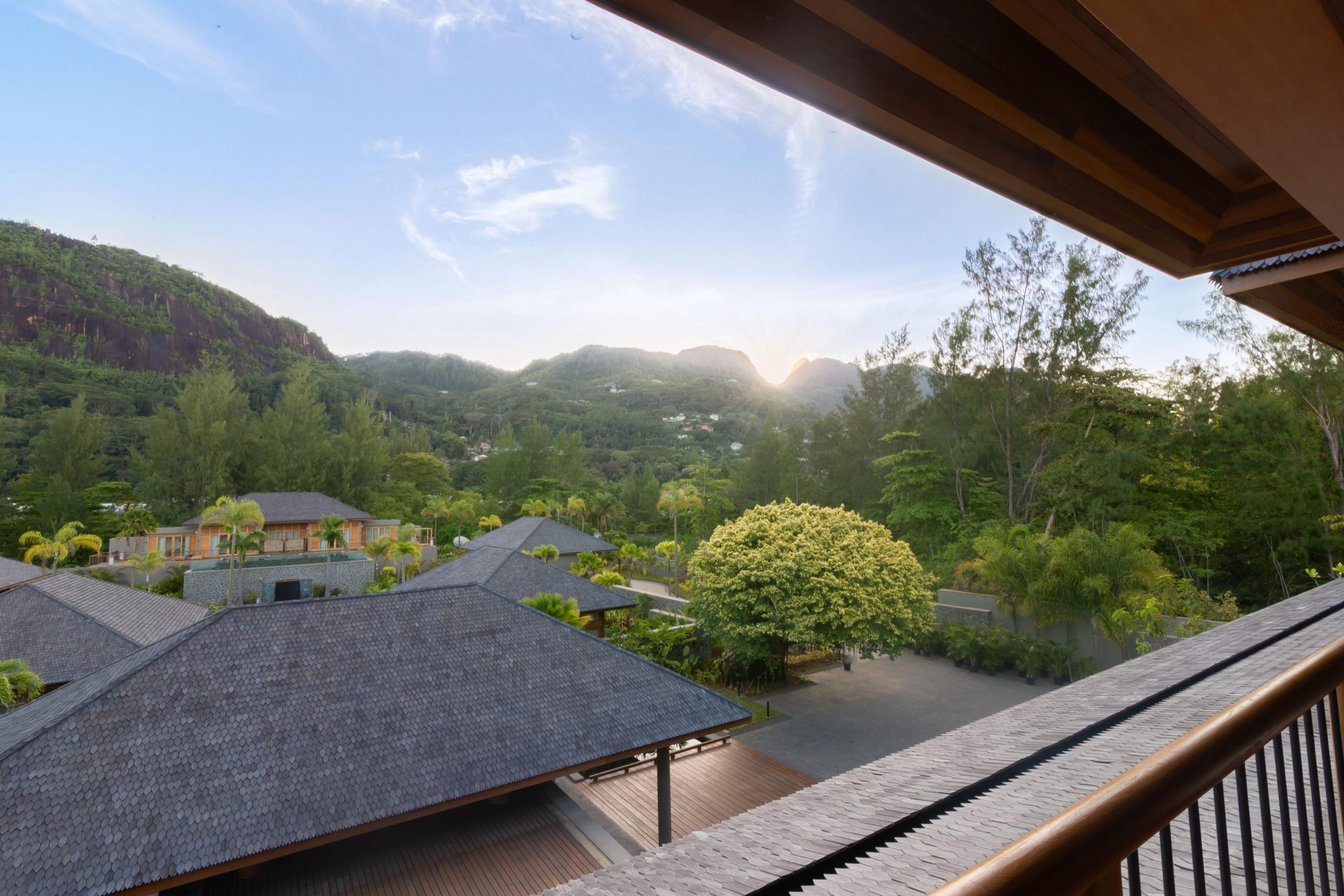 A view from a balcony over tiled rooftops to forested hills, with the sun rising behind a mountain ridge.