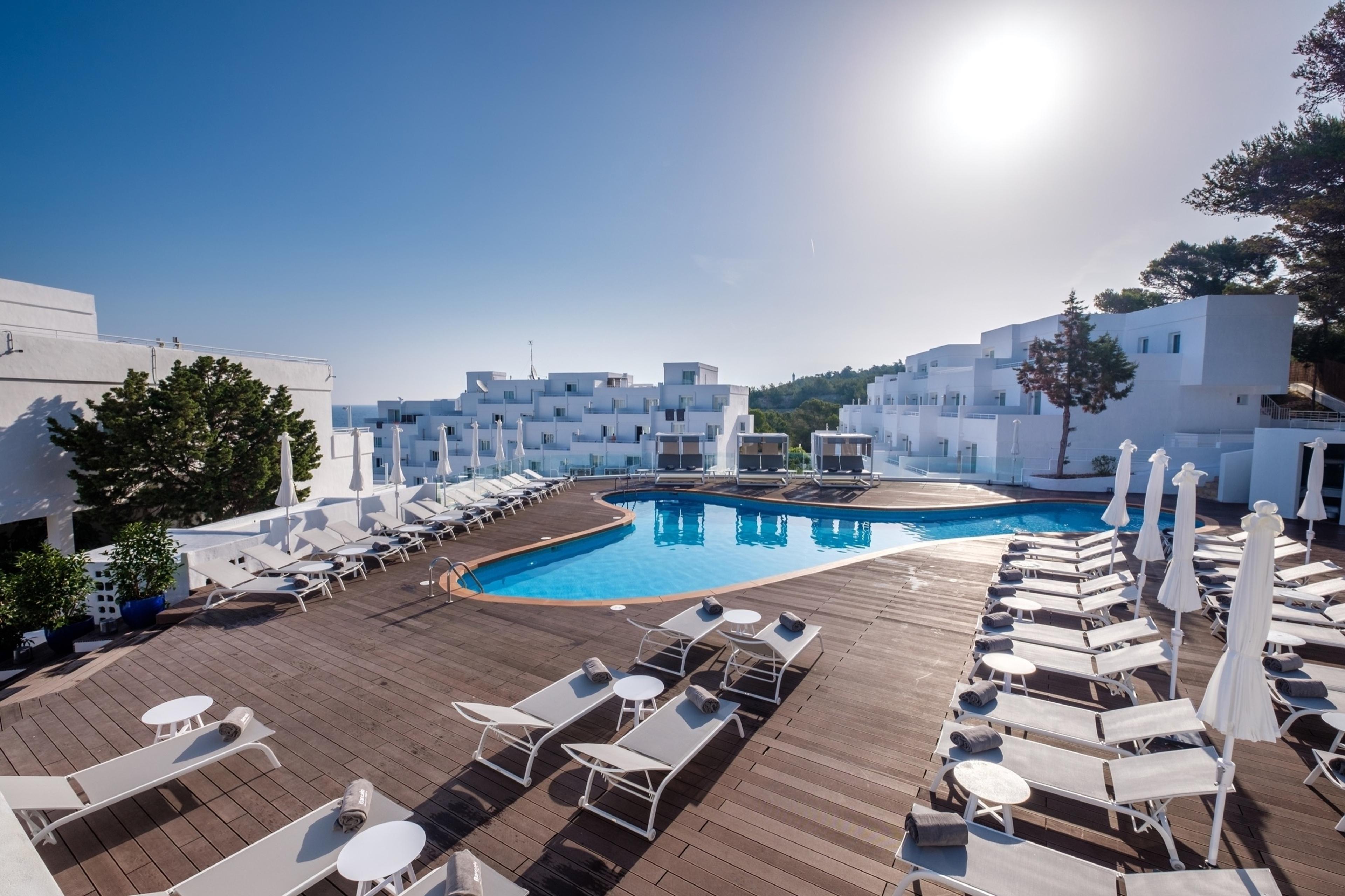 A curved swimming pool on a wooden sun deck surrounded by white sun loungers and umbrellas, with the hotel's white balconied buildings and pine trees around it under a bright sky.