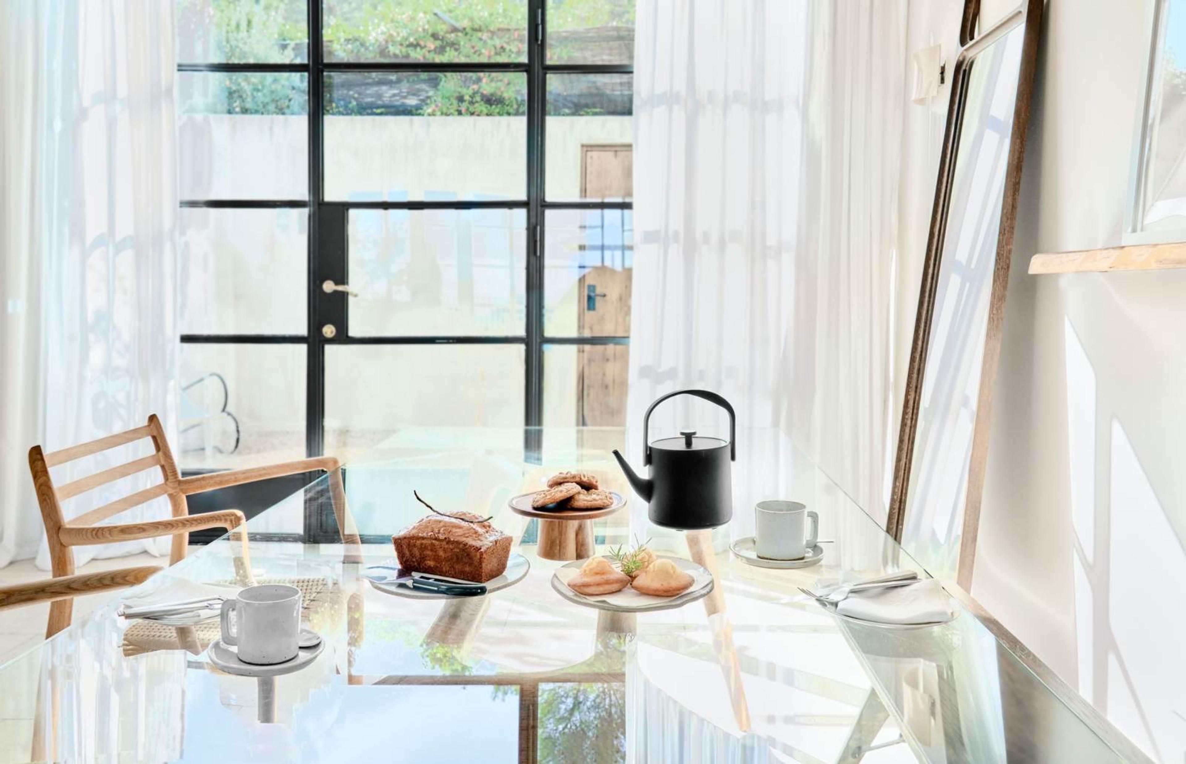 A glass-topped table set with banana bread, a stack of cookies, pastries, and a black teapot with white cups, beside glass doors hung with sheer curtains.