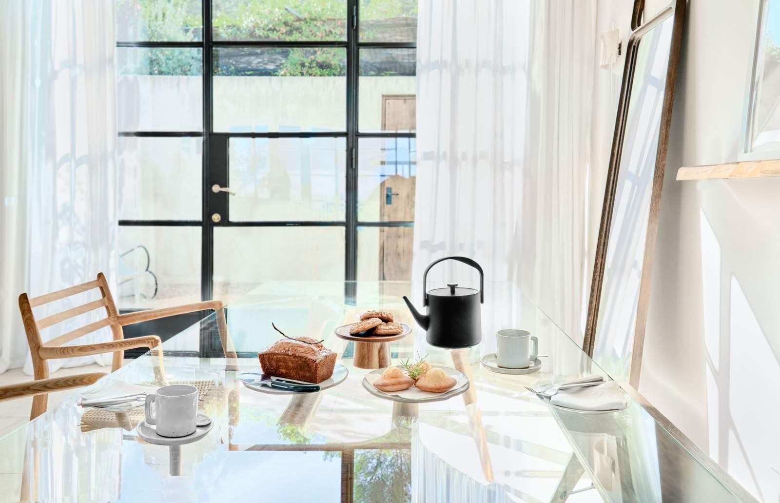 A glass-topped table set with banana bread, a stack of cookies, pastries, and a black teapot with white cups, beside glass doors hung with sheer curtains.