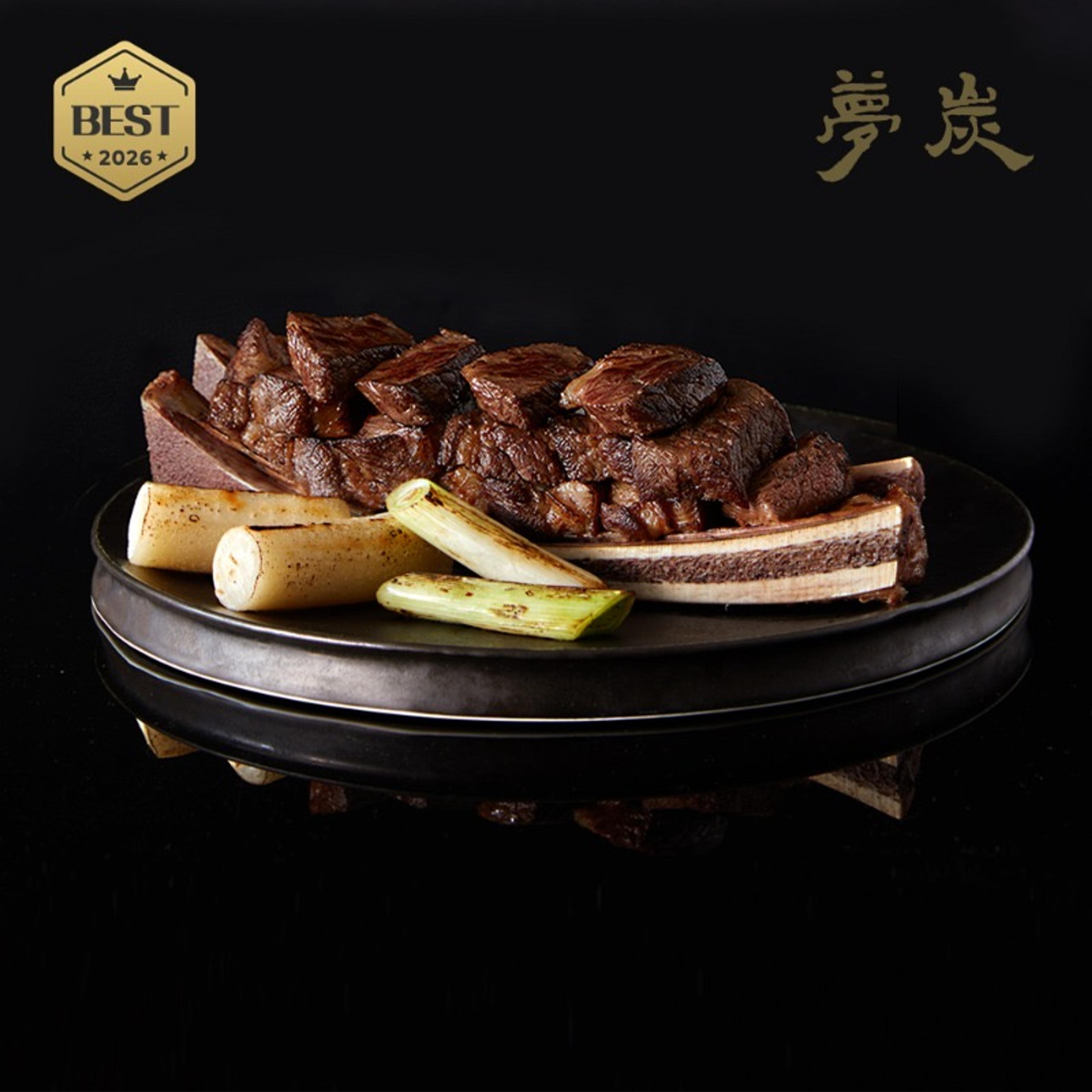 Straw-fire grilled beef short rib, cut from the bone, with charred spring onions on a black plate.