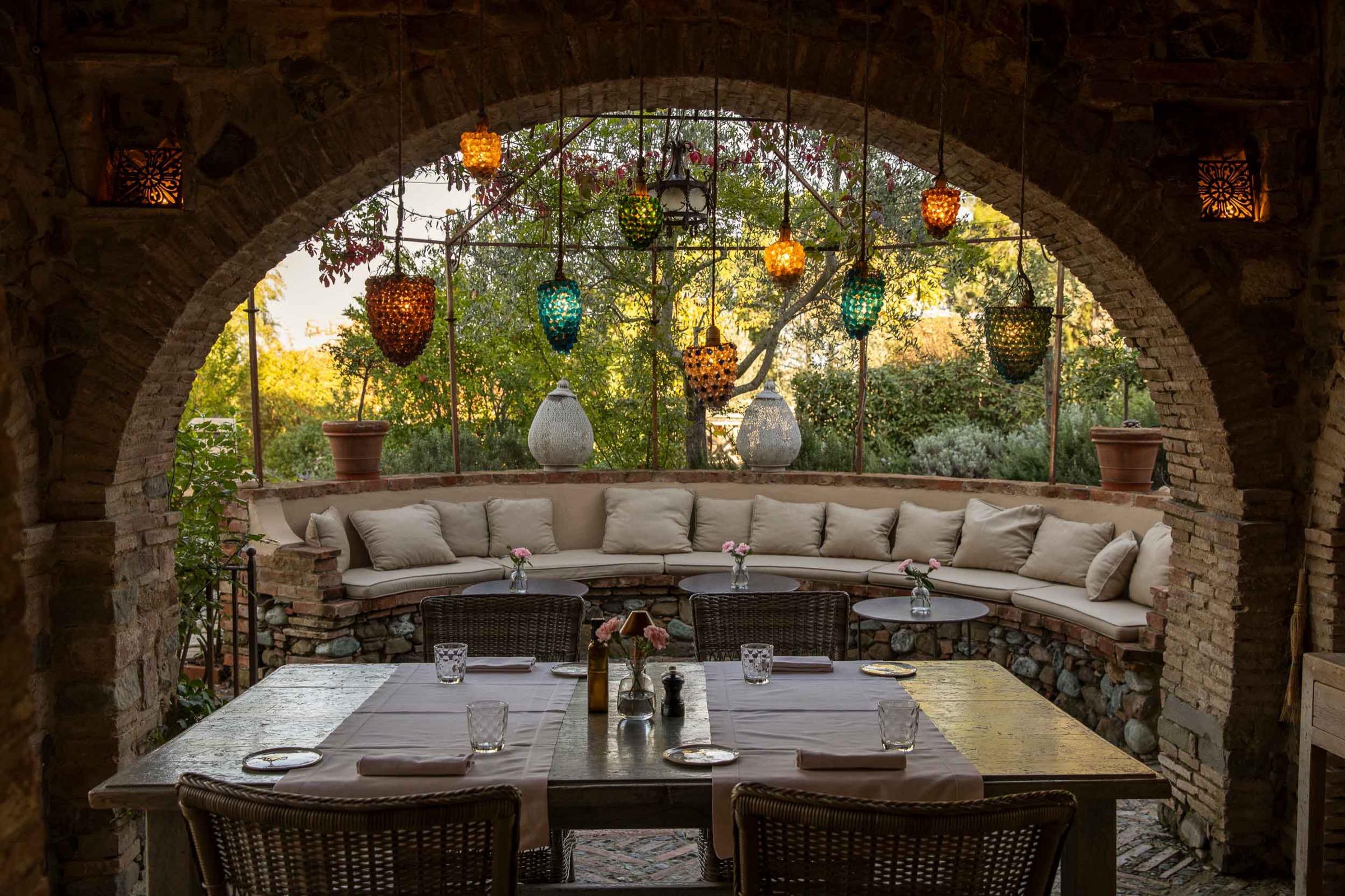 A table set for dinner under a stone pergola strung with coloured glass lanterns, with a curved cushioned bench behind.