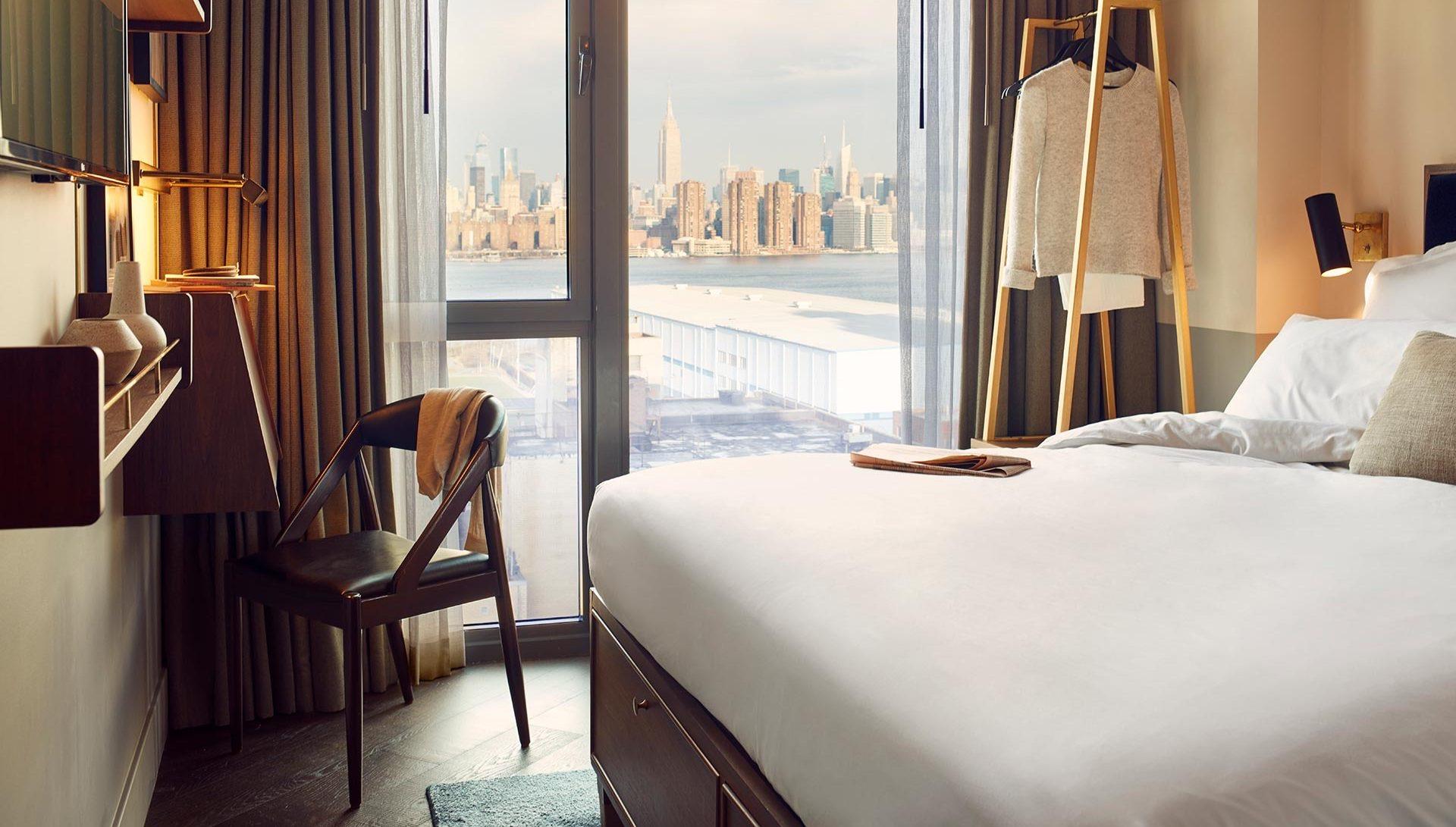 a chair by a floor-to-ceiling window showing the Manhattan skyline across the East River, with a clothing rail and the edge of a bed in the room