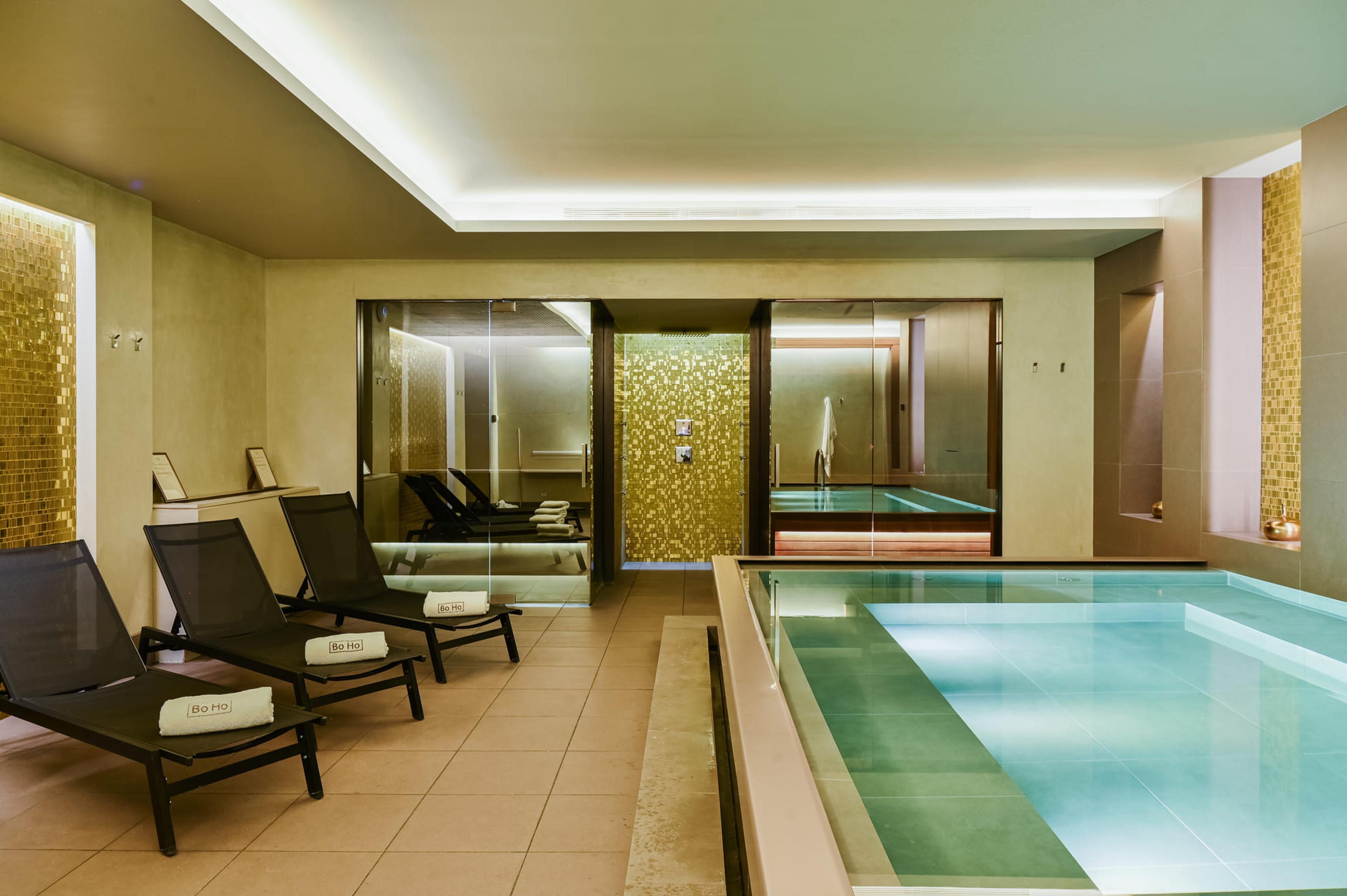 An indoor pool with pale turquoise water, black loungers on tiles beside it, and glass-walled rooms behind.