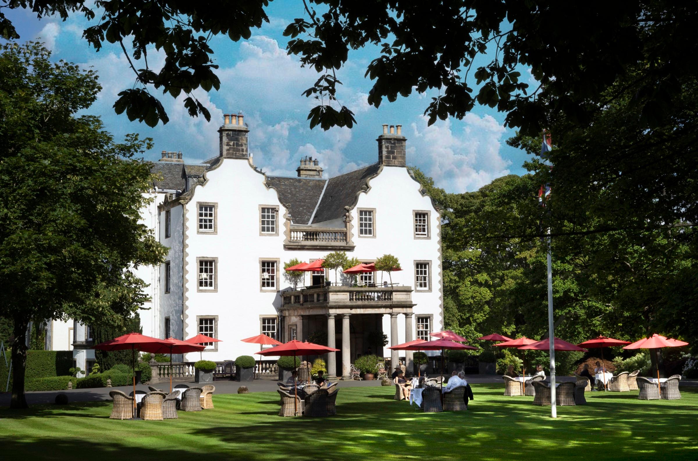 the white harled facade of Prestonfield House under a blue sky, with red umbrellas and wicker chairs set out on the lawn in front