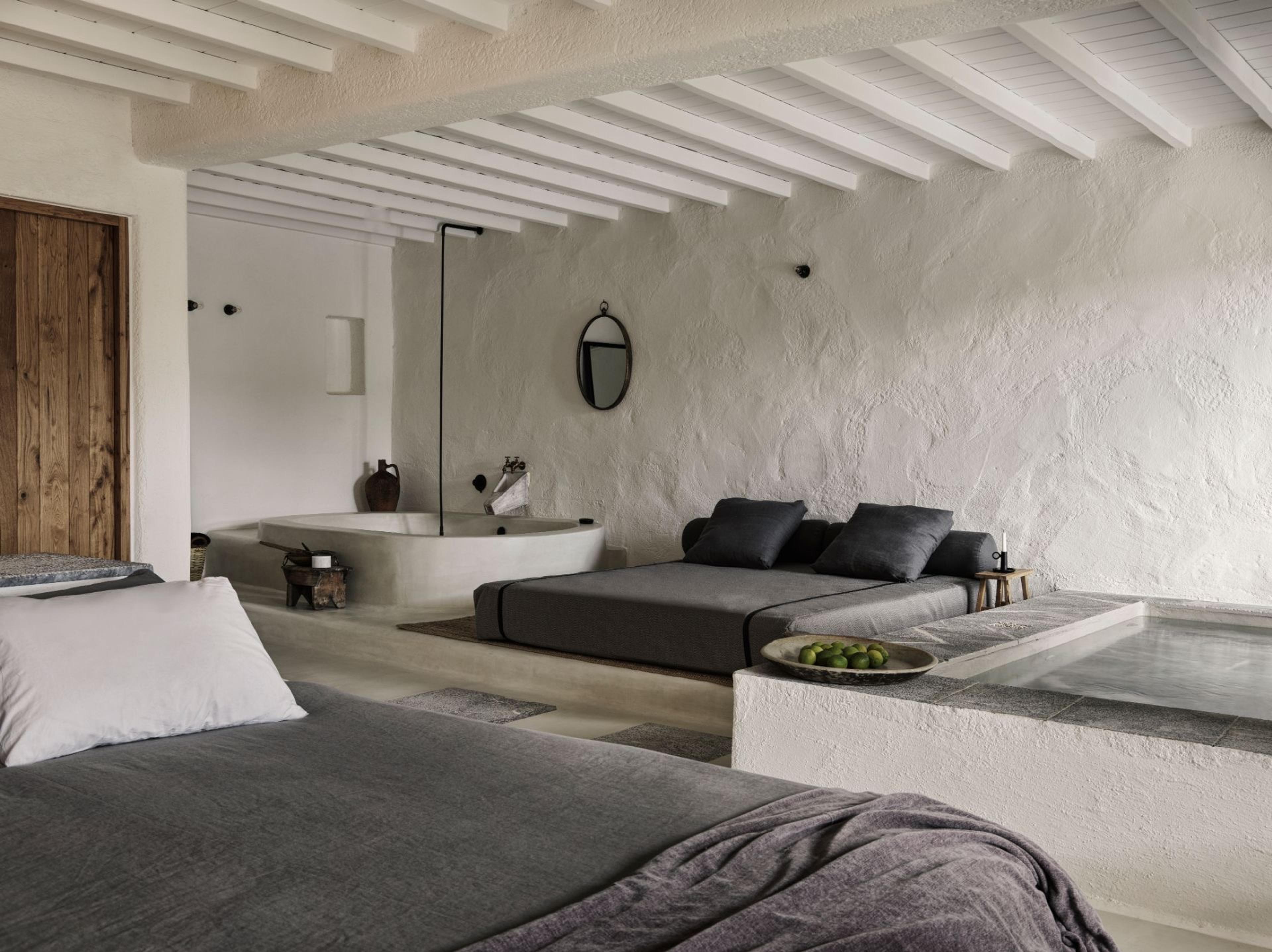 A bedroom with a dark beamed ceiling has two beds in grey linen facing a stone plunge tub and a built-in pool set into the floor, a bowl of green apples on the ledge between them.