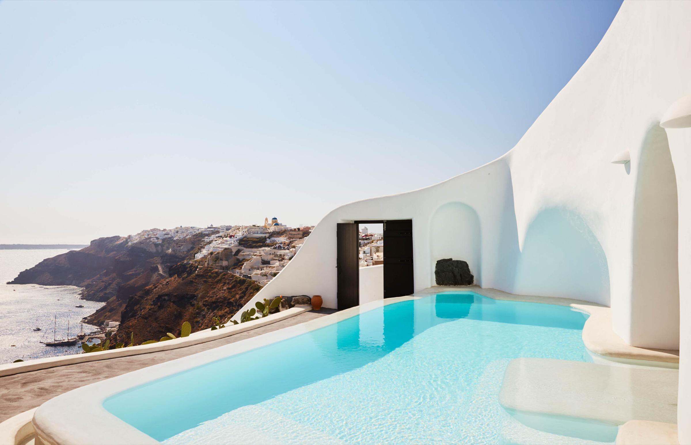 A curved whitewashed building beside an infinity pool, with cactus plants on the terrace and a clifftop village in the distance.