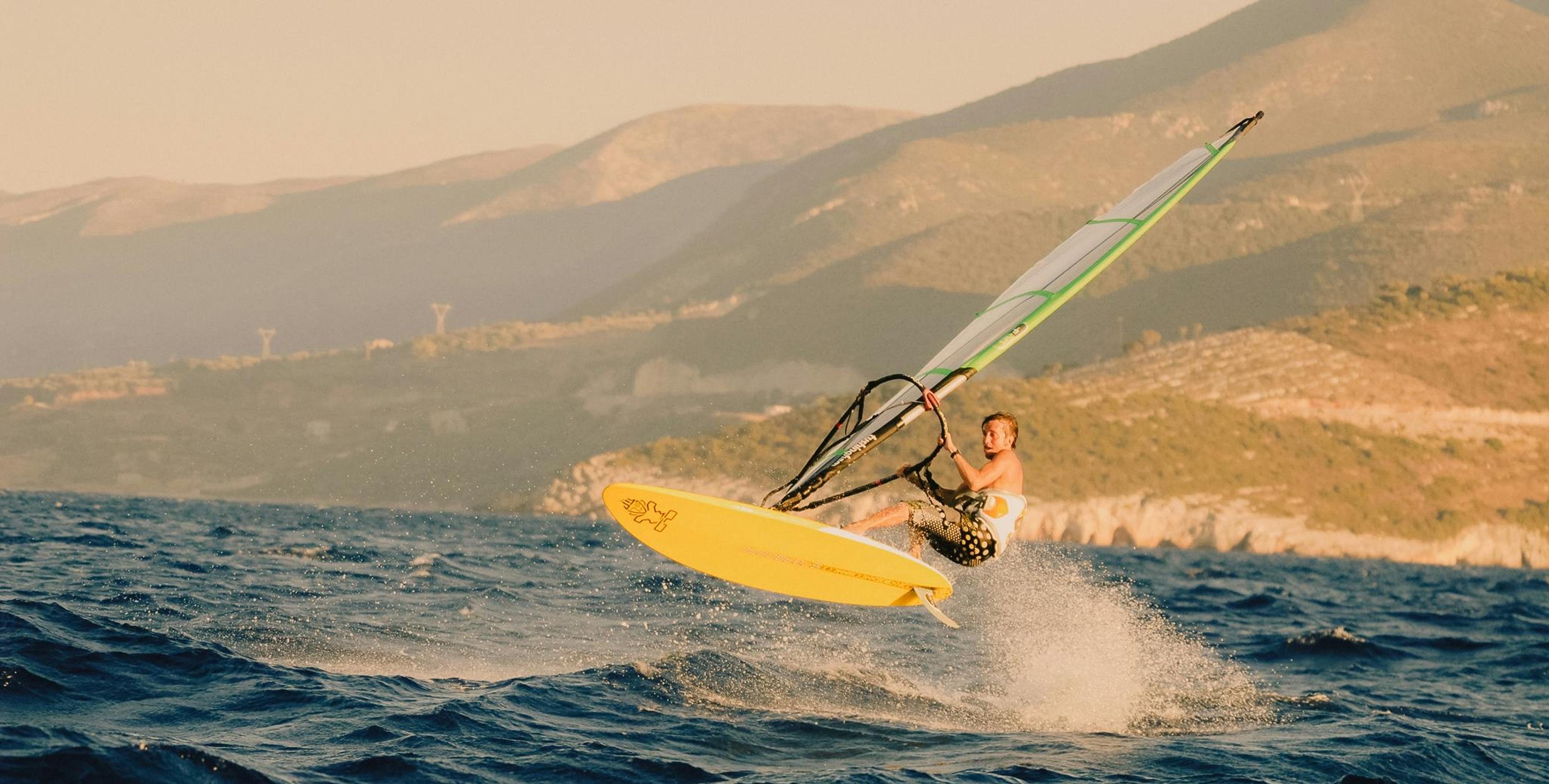 A windsurfer airborne off a yellow board, sail angled against a hazy mountain coastline.