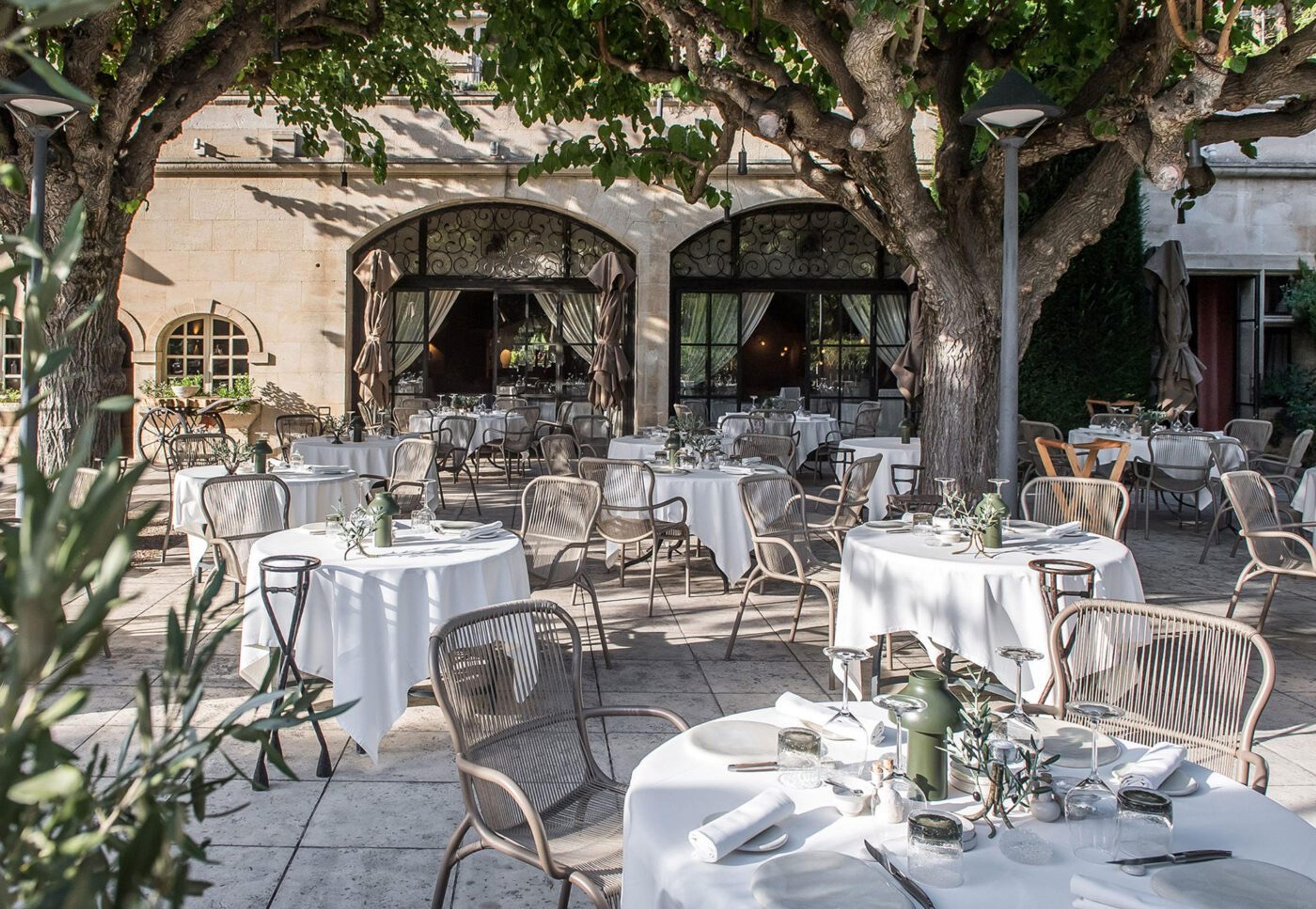 A restaurant terrace shaded by two large plane trees, with white-clothed tables set for lunch in front of the stone hotel building.