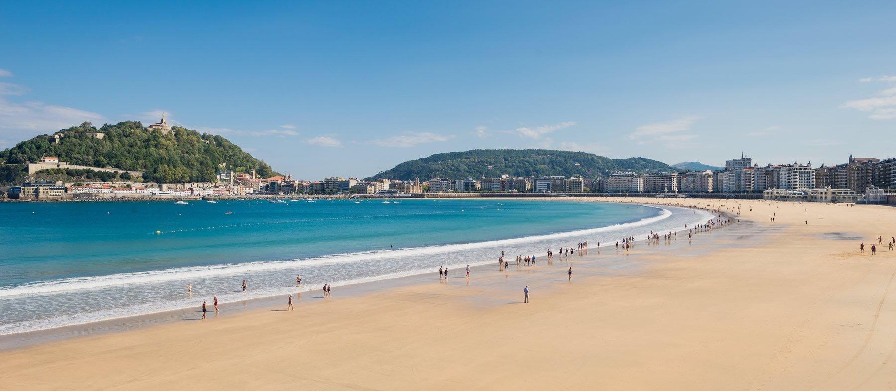 A broad sandy beach curving around a bay, with a tree covered hill and a hilltop building at one end, and small groups of people walking near the water.