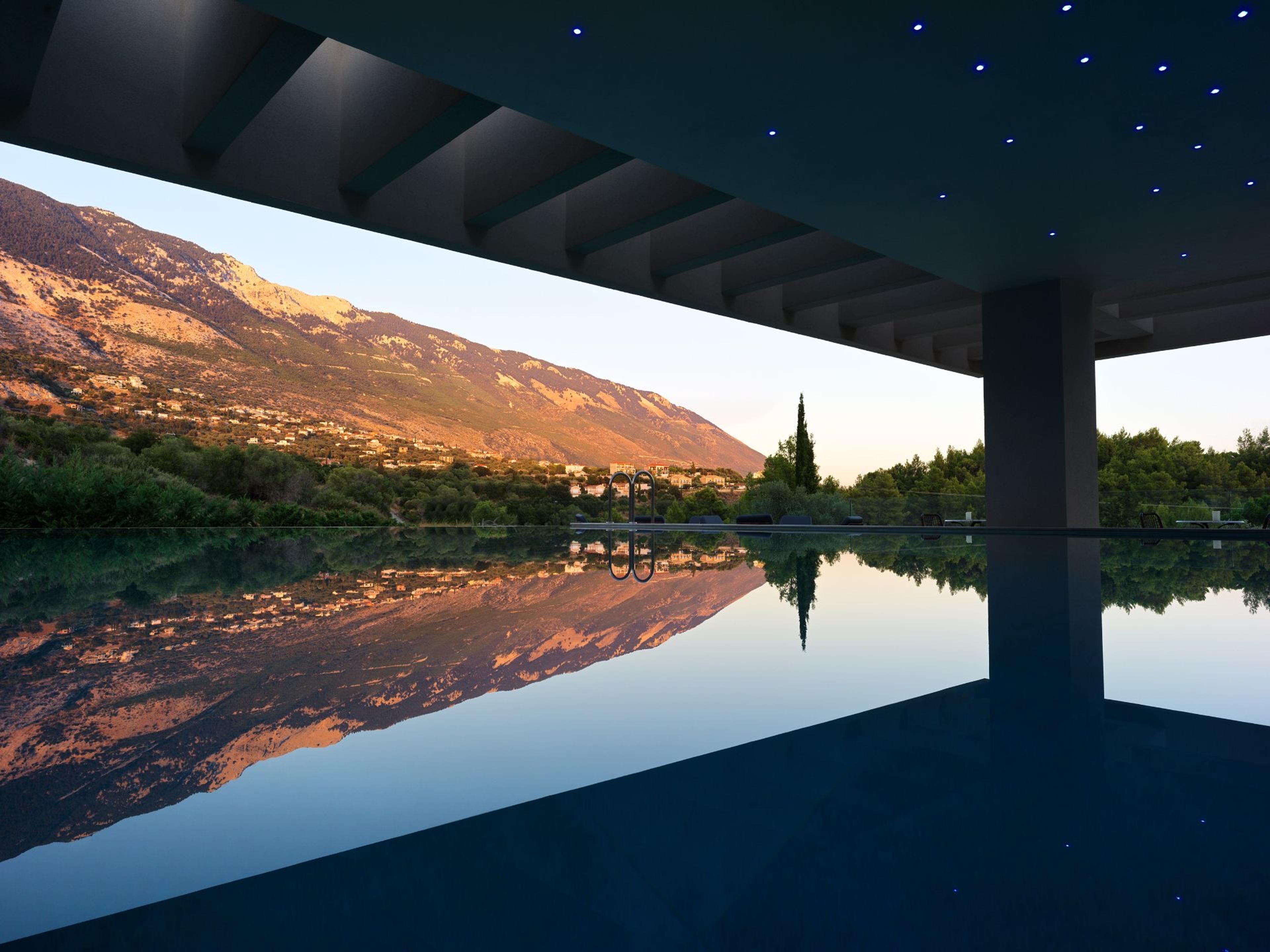 A covered poolside terrace at dusk with a star-lit ceiling, overlooking an infinity pool reflecting a sunlit mountain.