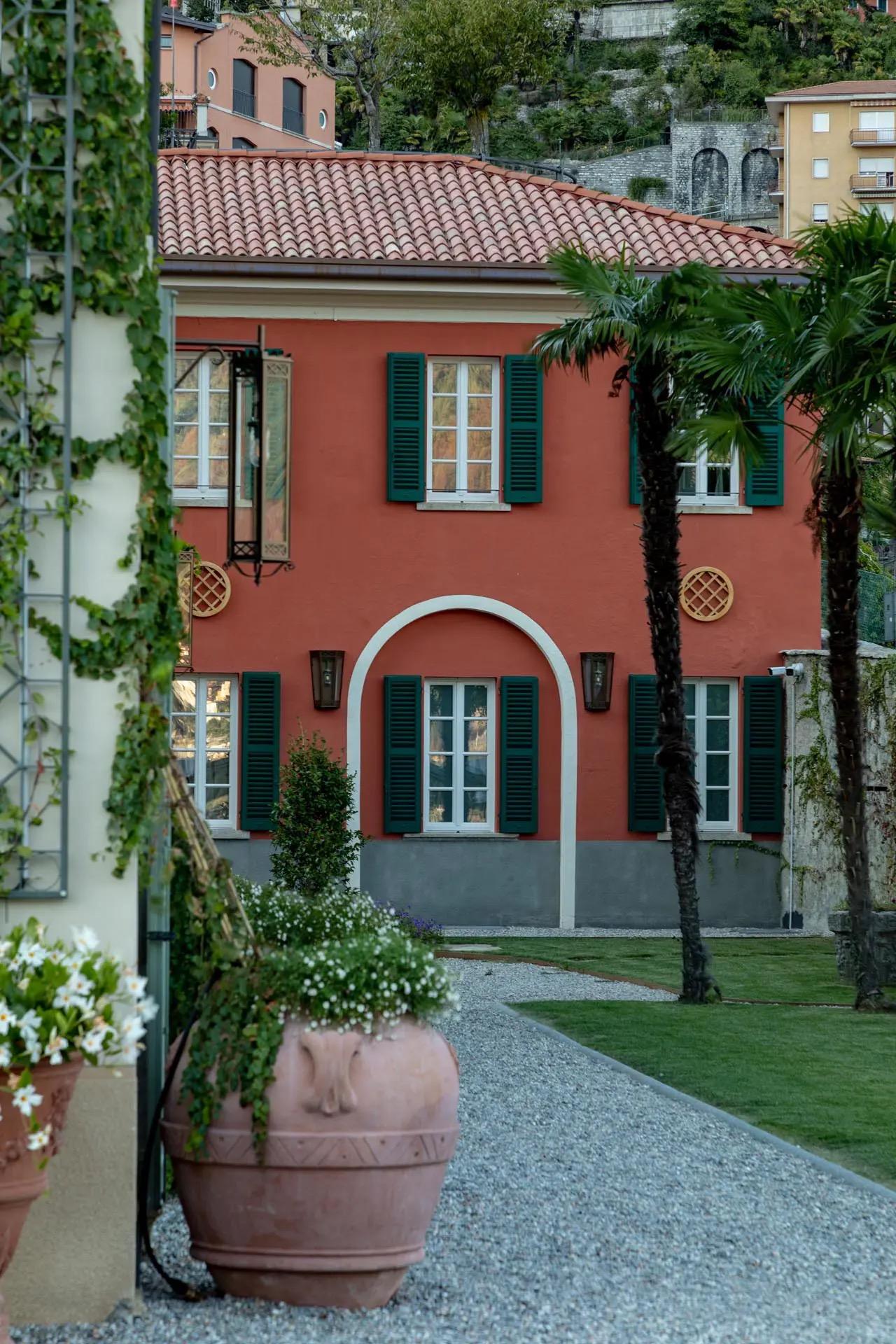A terracotta-colored villa with green shutters and an arched doorway, seen past potted plants and two palm trees on a gravel path.