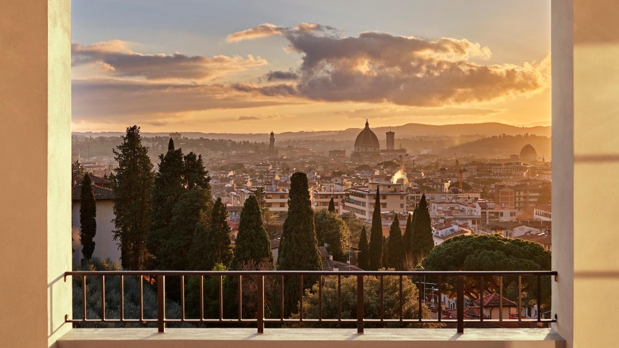 a balcony framed between two stone pillars, looking across cypress trees to the Florence skyline and the Duomo at sunset
