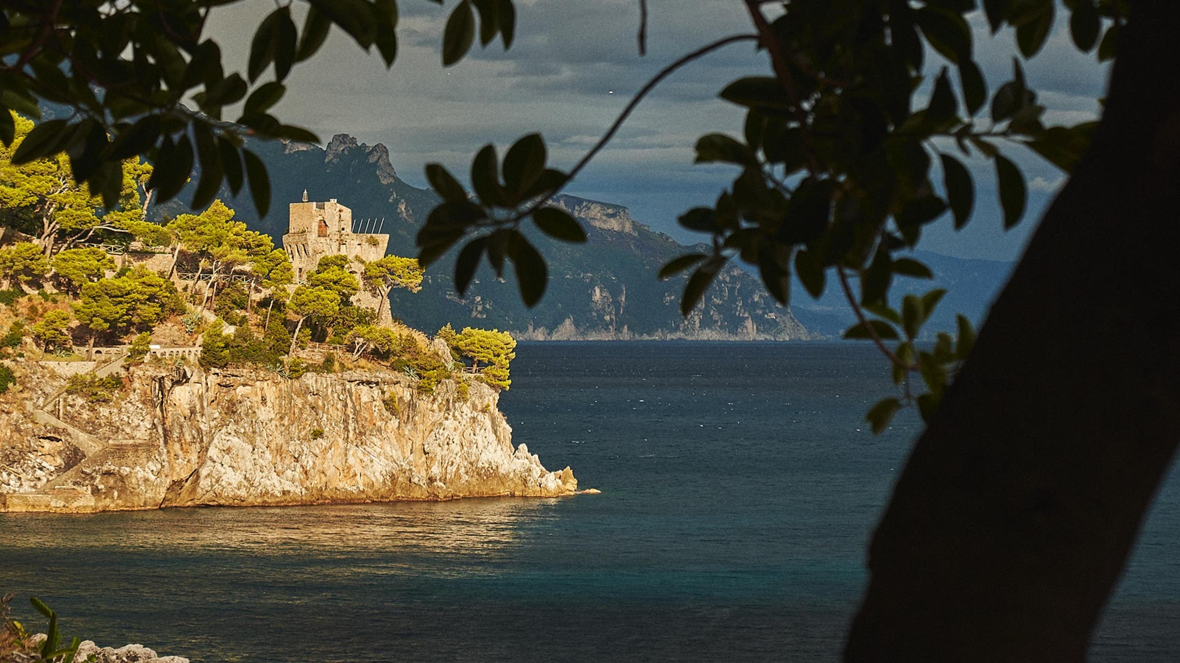 A stone watchtower on a wooded headland above the sea, framed by dark tree branches in the foreground, with mountains fading into haze beyond.