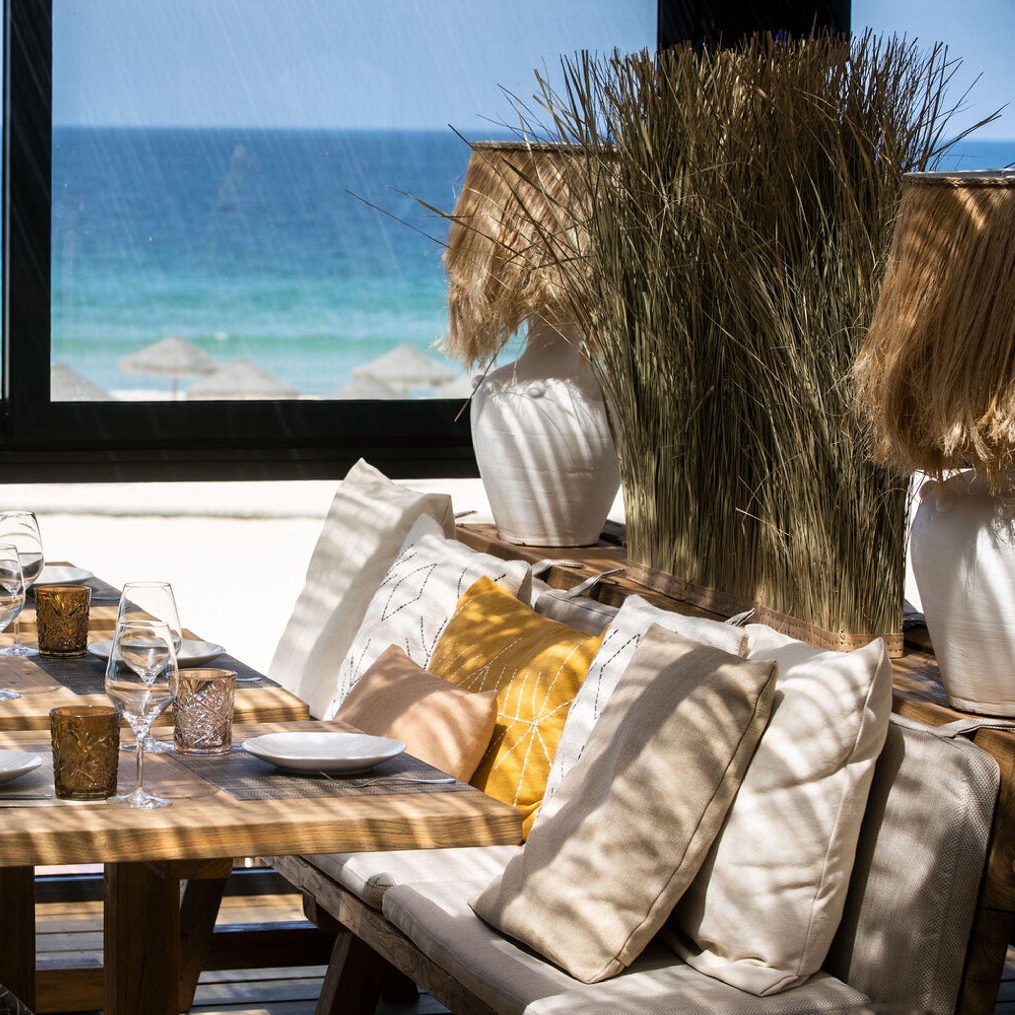 Cushioned banquette seating and set tables under straw parasols, the sea beyond the glass