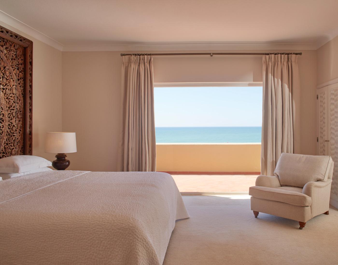 A bedroom with a carved wooden headboard, a made bed, a cream armchair and a curtained doorway open to a sea view.
