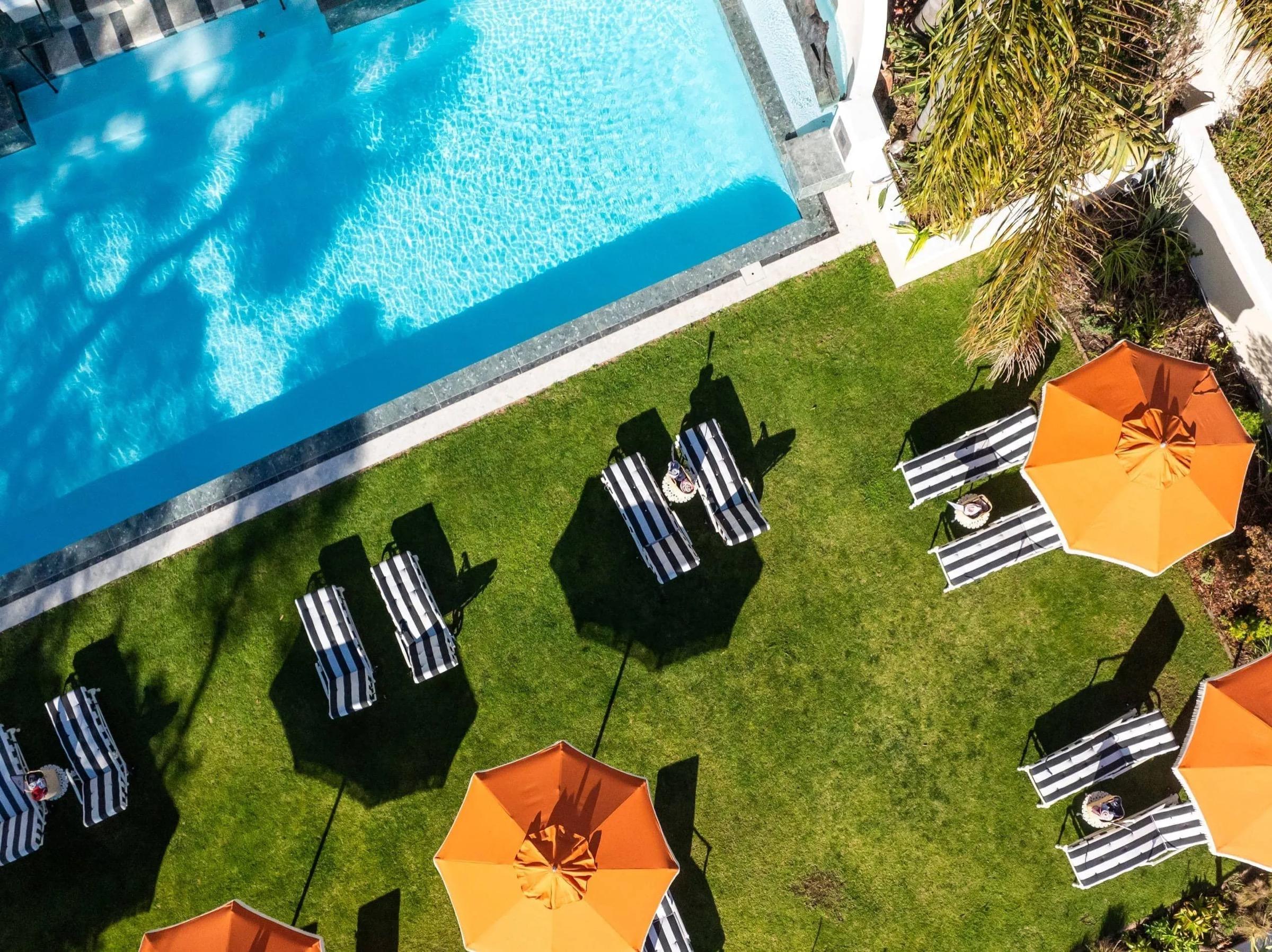 An aerial view of a turquoise swimming pool beside a lawn with striped sun loungers and orange parasols.