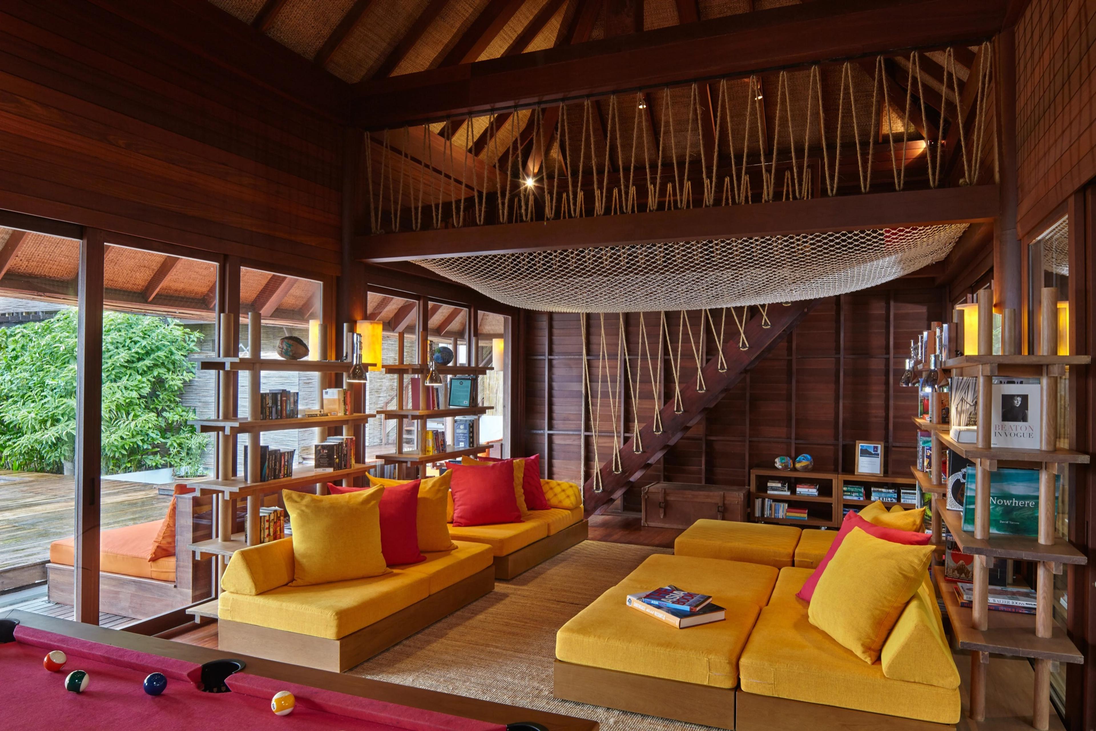 A game room with a pool table in the foreground and built-in yellow sofas under a high thatched ceiling, a rope-strung hammock slung above a staircase and a wall of books to the side.