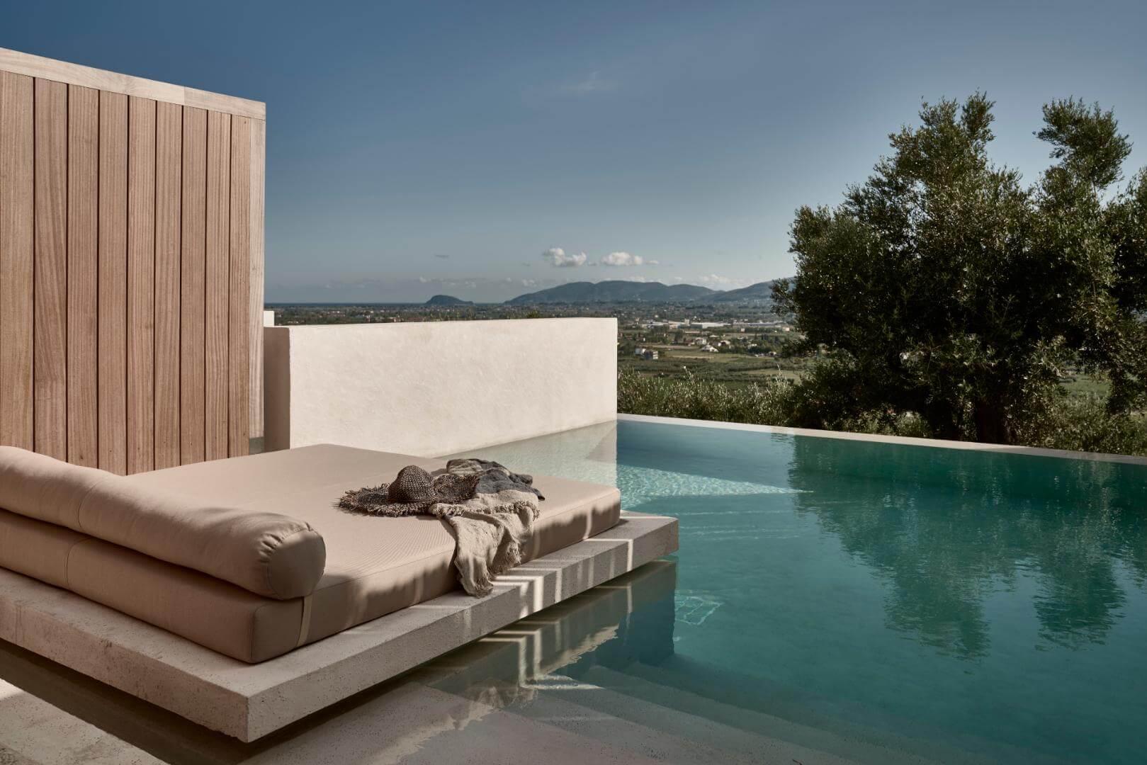 A built-in daybed with a folded blanket sits beside an infinity pool, overlooking hills and a distant coastline.