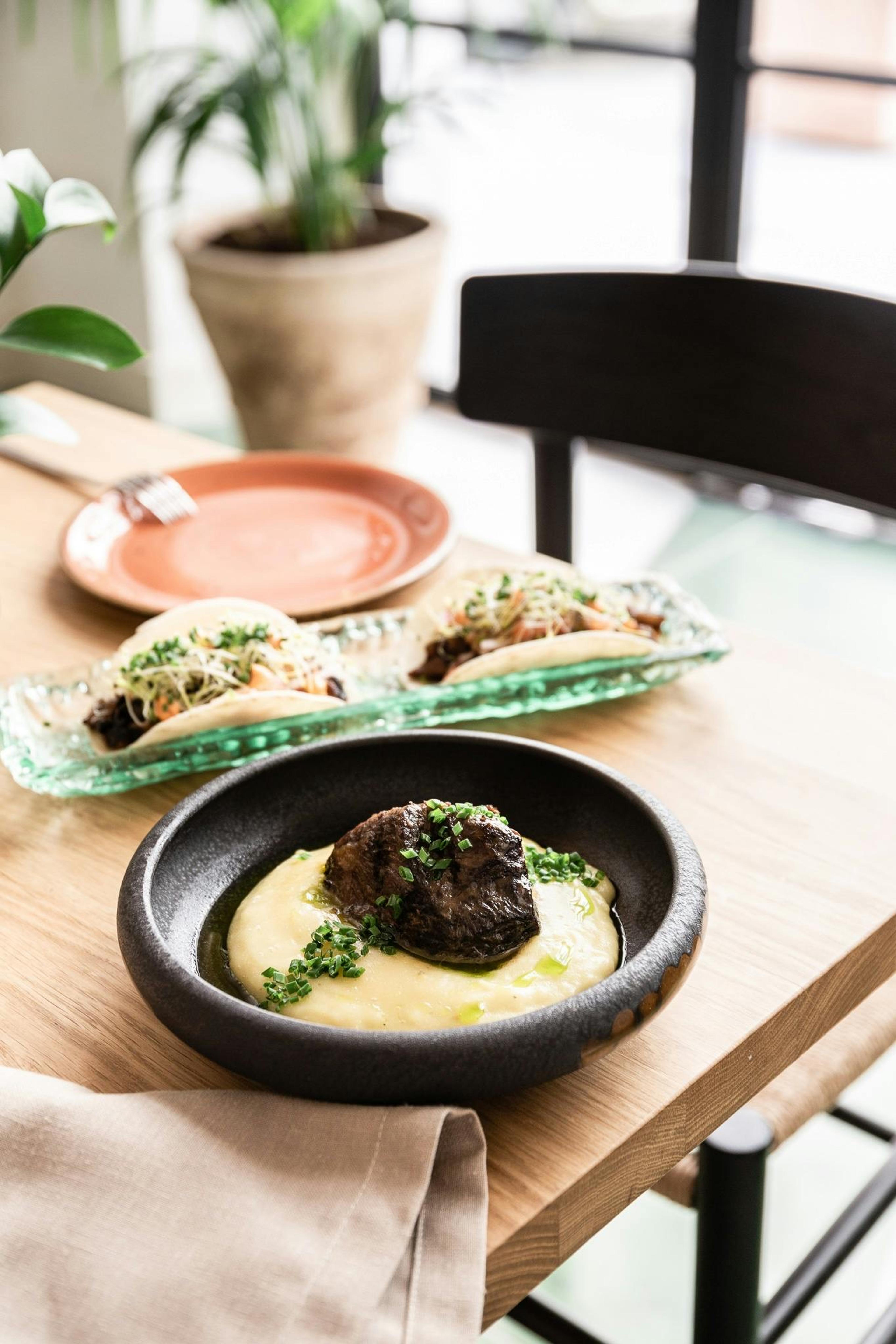 A seared piece of meat on a bed of pureed potato in a black bowl, with two folded tacos and an empty terracotta plate on a wooden table.