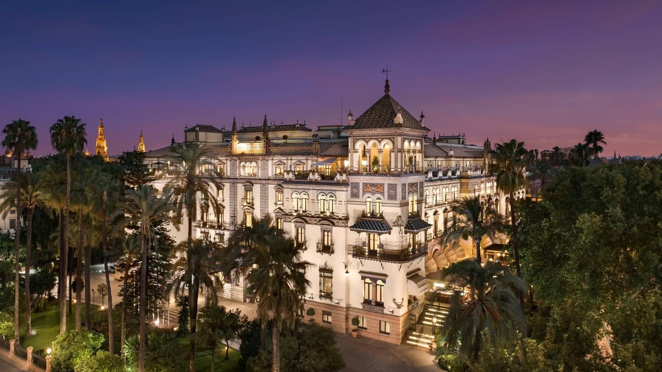 A grand white and ochre hotel building lit at dusk, with a central tiled tower, palm trees in the foreground and a cathedral bell tower visible in the distance.