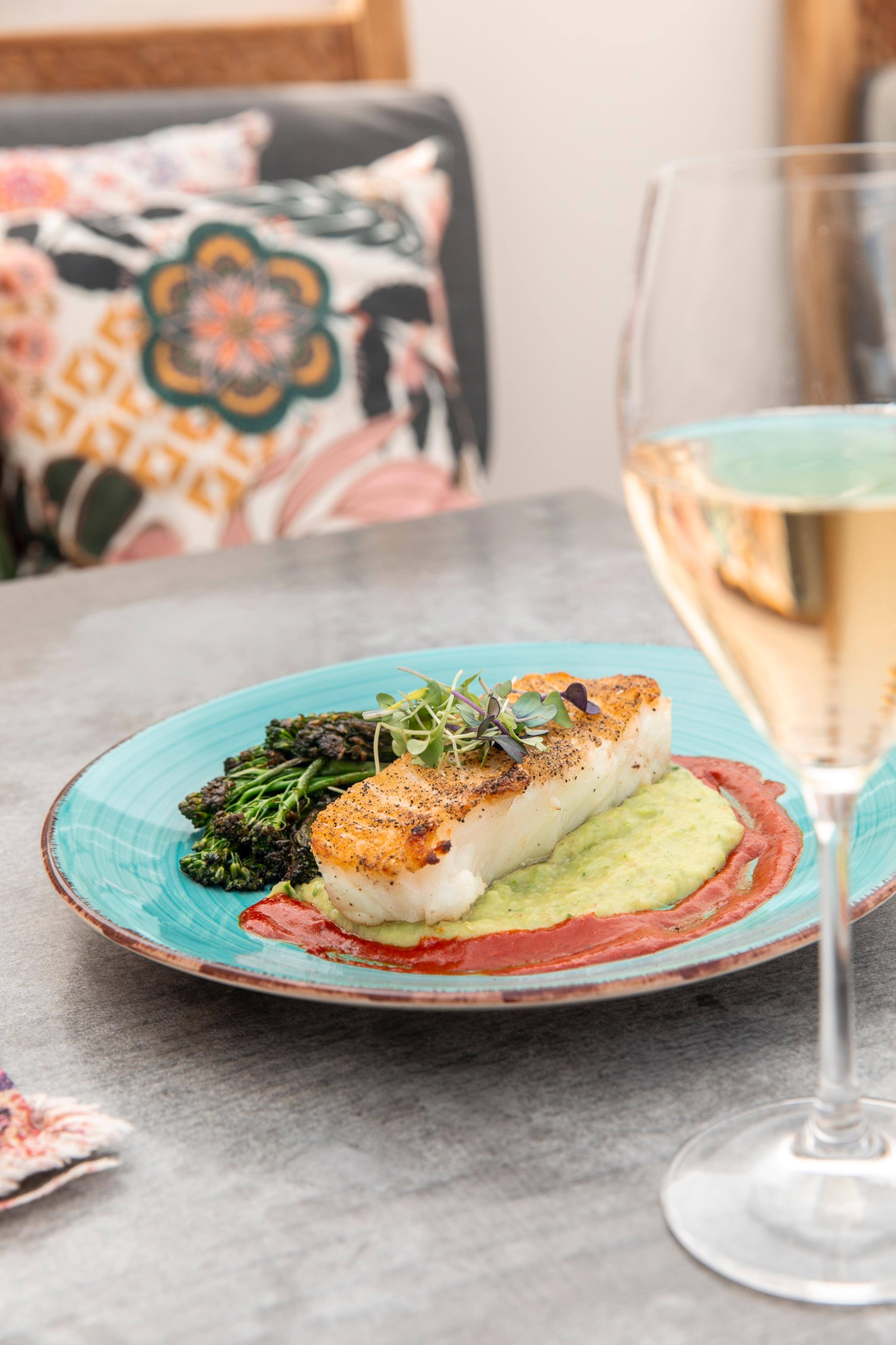 A plated fillet of fish with charred broccolini on a green sauce, beside a glass of white wine.