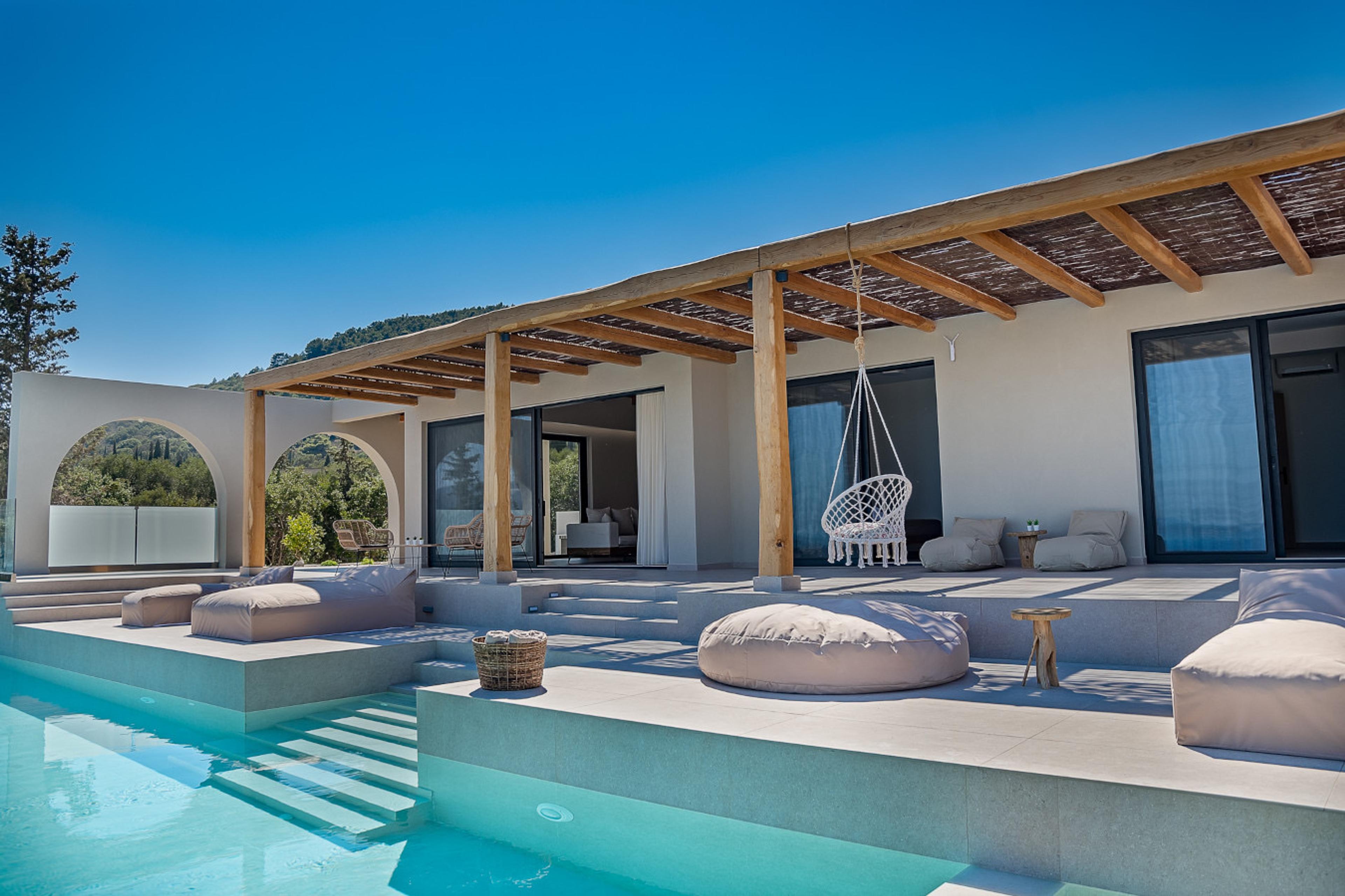 A white villa with a wooden pergola and arched openings, seen across an infinity pool with loungers and a hanging swing chair on the terrace.