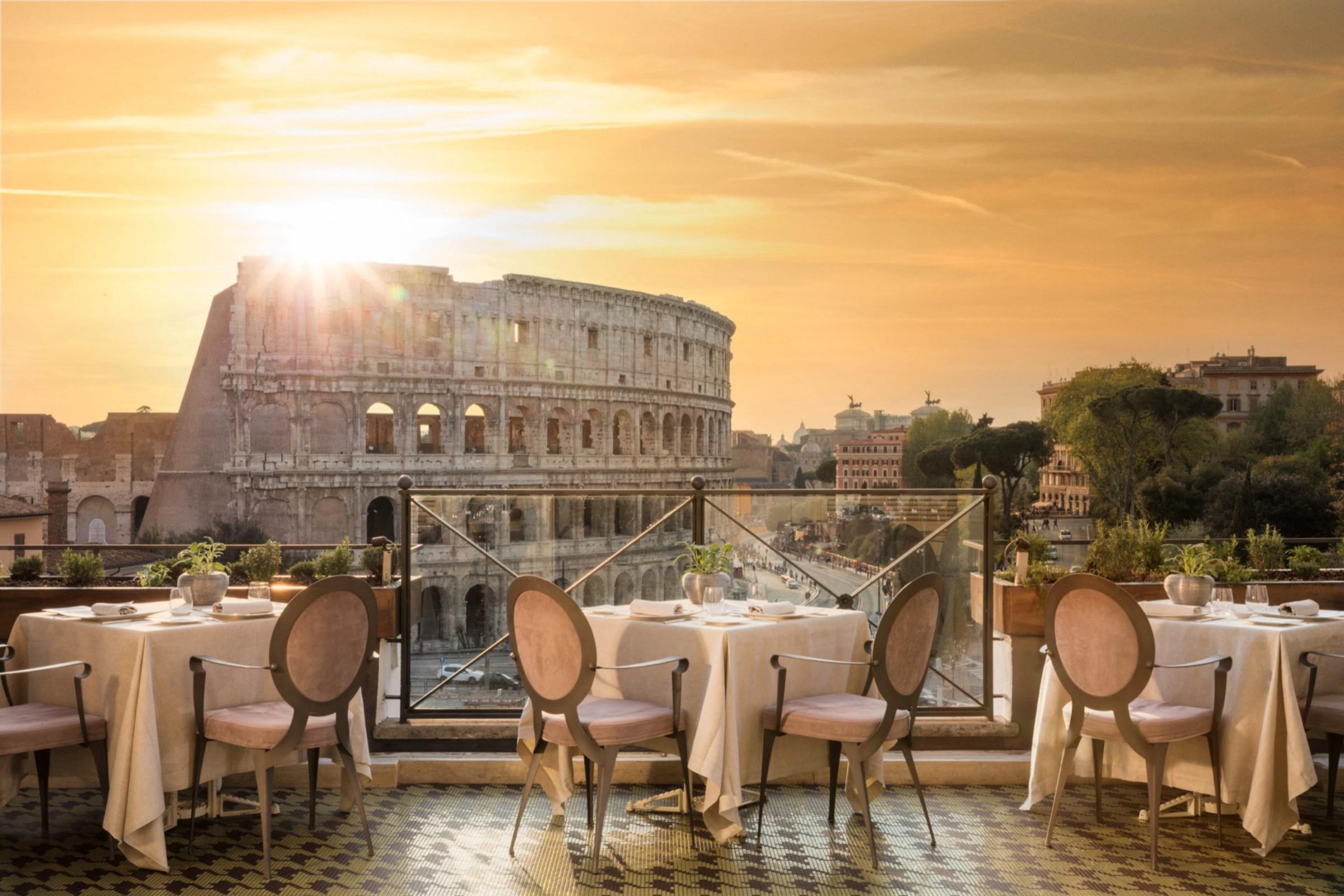A rooftop terrace laid for dinner at sunset, with cream tablecloths and pink-cushioned chairs, and the Colosseum lit behind the railing.