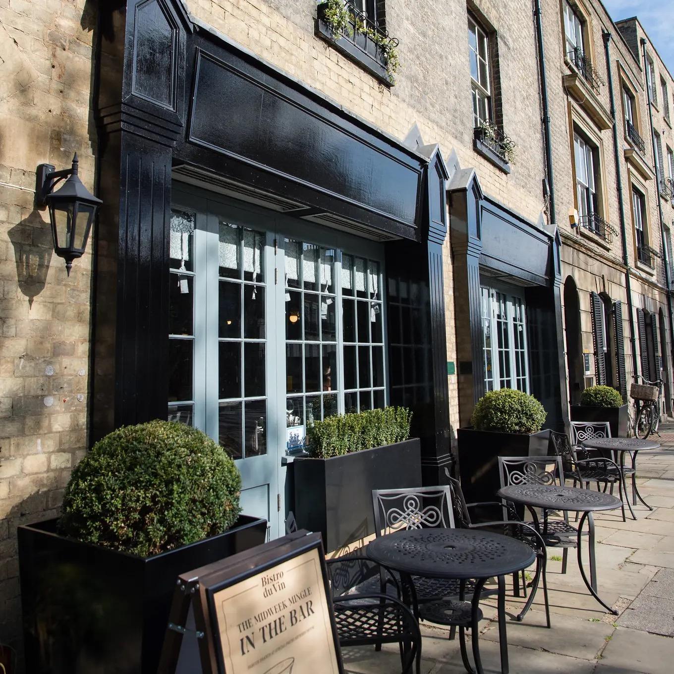 The front of a bistro on a paved street, with a black painted fascia, pale blue-grey doors, clipped box hedges in tall planters, and small metal tables and chairs set out on the pavement.