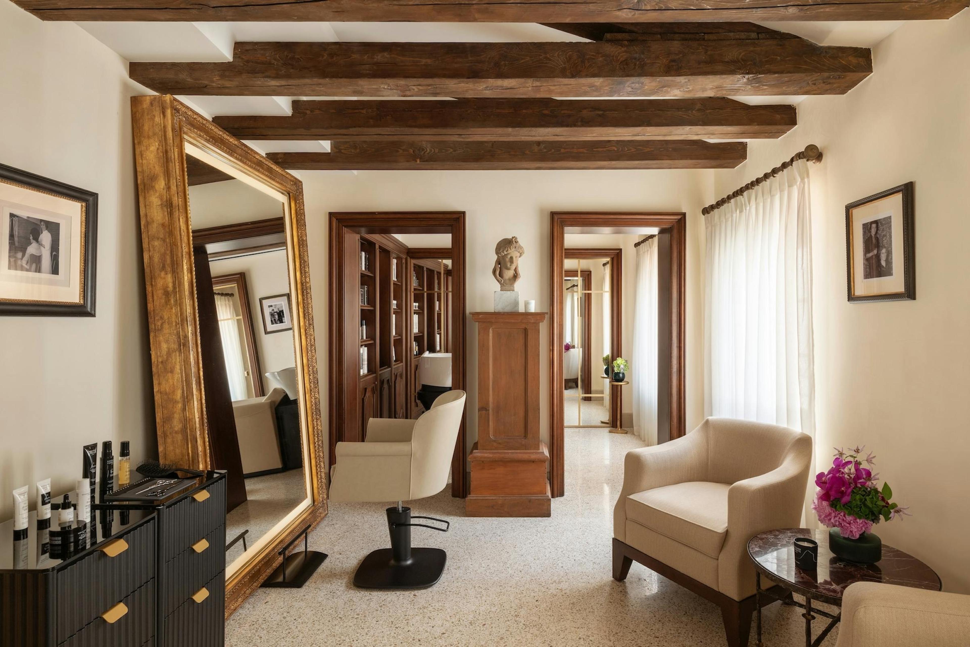 A beamed room with a large gilt-framed mirror, a salon chair, a stone pedestal with a bust, cream armchairs, and a doorway through to another mirrored room.