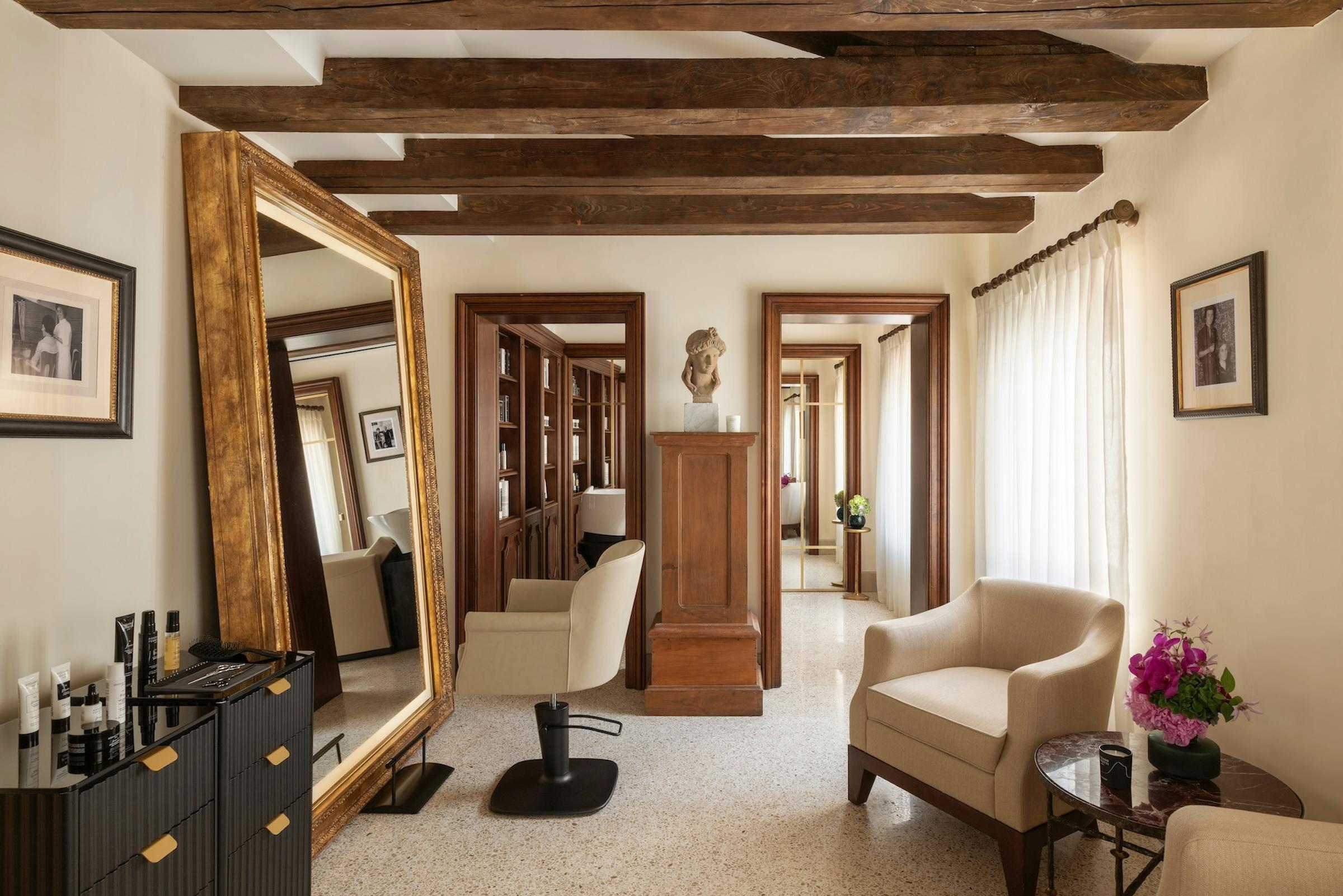 A beamed room with a large gilt-framed mirror, a salon chair, a stone pedestal with a bust, cream armchairs, and a doorway through to another mirrored room.