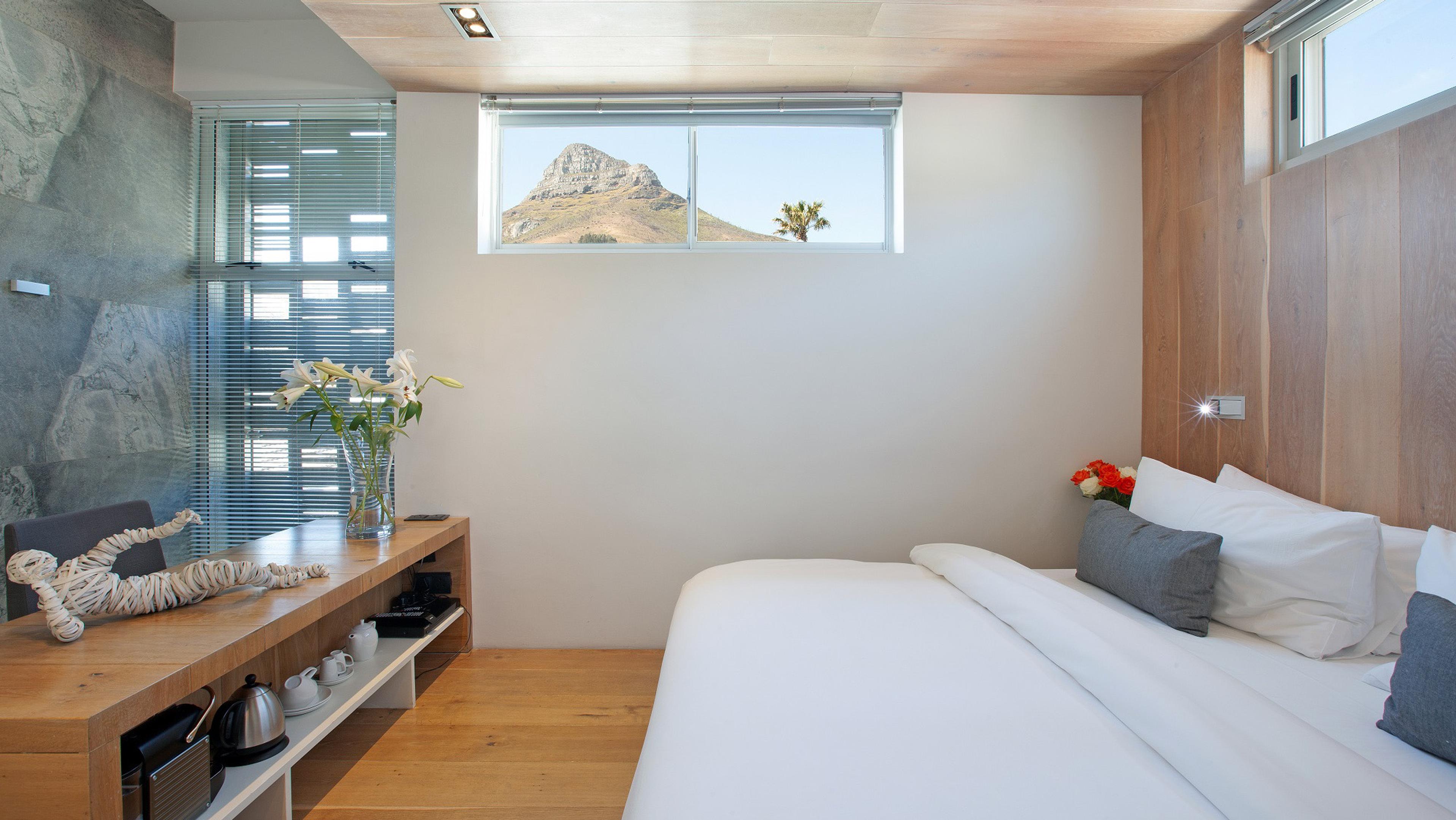 A made-up bed beneath a small window framing Lion's Head mountain, with a vase of flowers and a woven ornament on the sideboard.