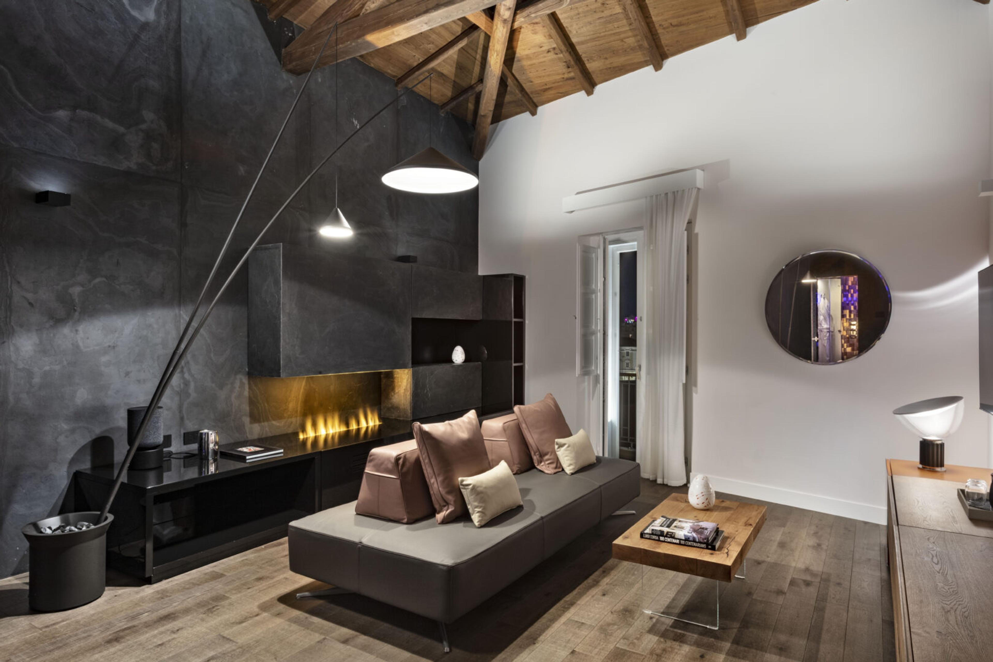 A living room at night with a stone wall, a built-in fireplace, a grey sofa with cushions, a round mirror, and glass doors open to a lit balcony.