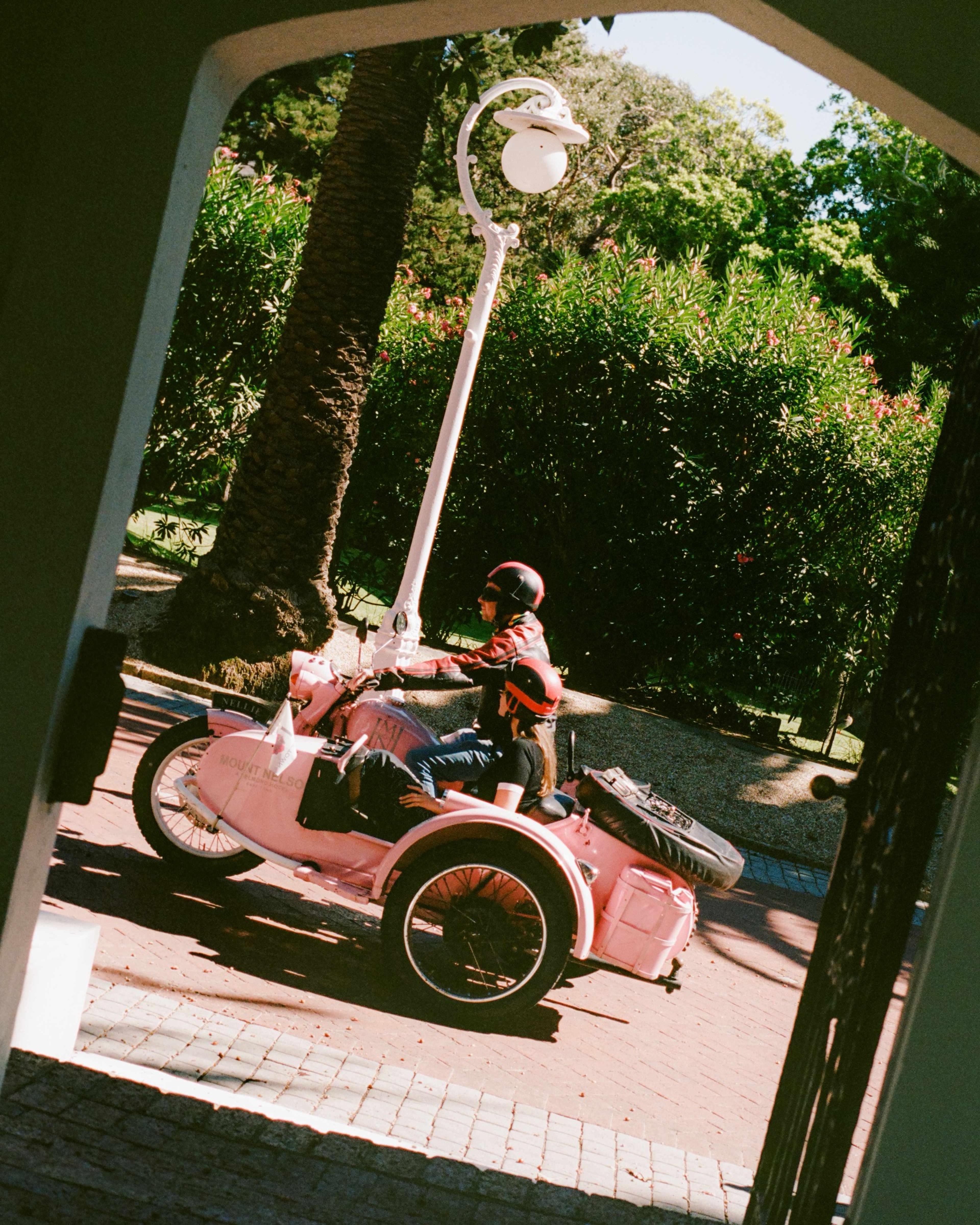 A pink sidecar motorcycle with two riders in helmets on a brick path under palm trees and oleanders, seen through an archway, with a white lamppost beside it.
