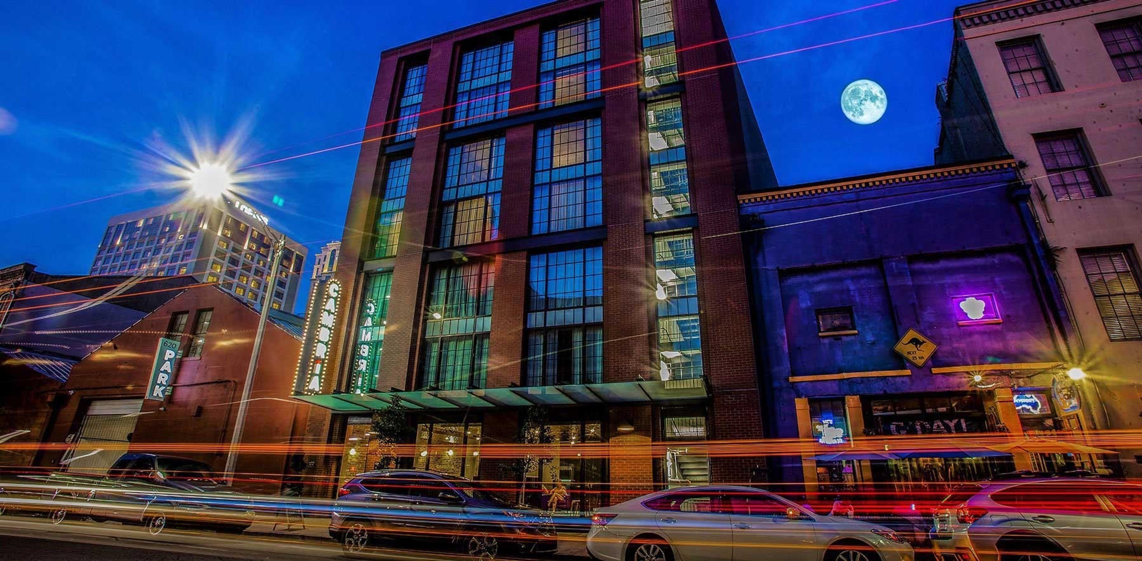 A hotel building lit up at night with an illuminated CAMBRIA sign, a full moon overhead, and light trails from passing traffic.