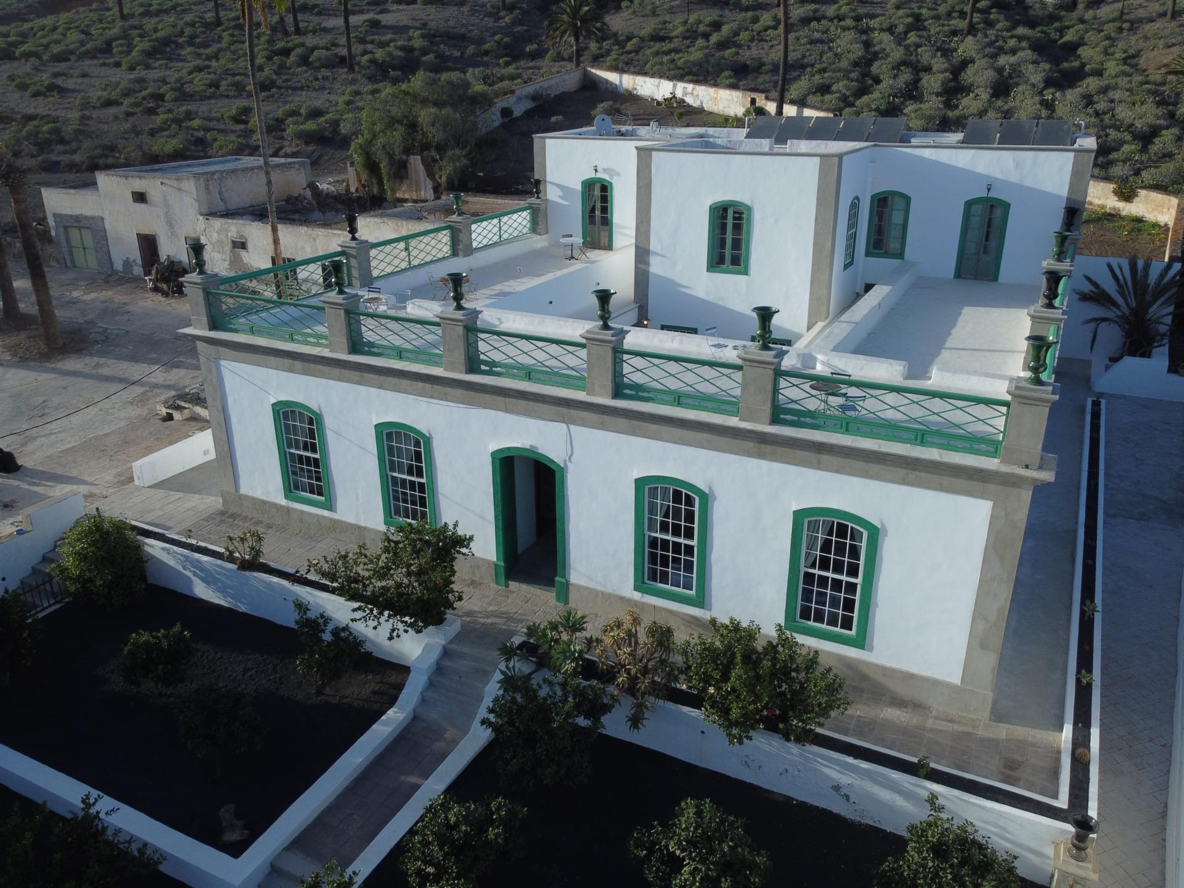 An aerial view of the whitewashed building with its green-railed roof terrace, surrounded by courtyards, outbuildings and scrubland.