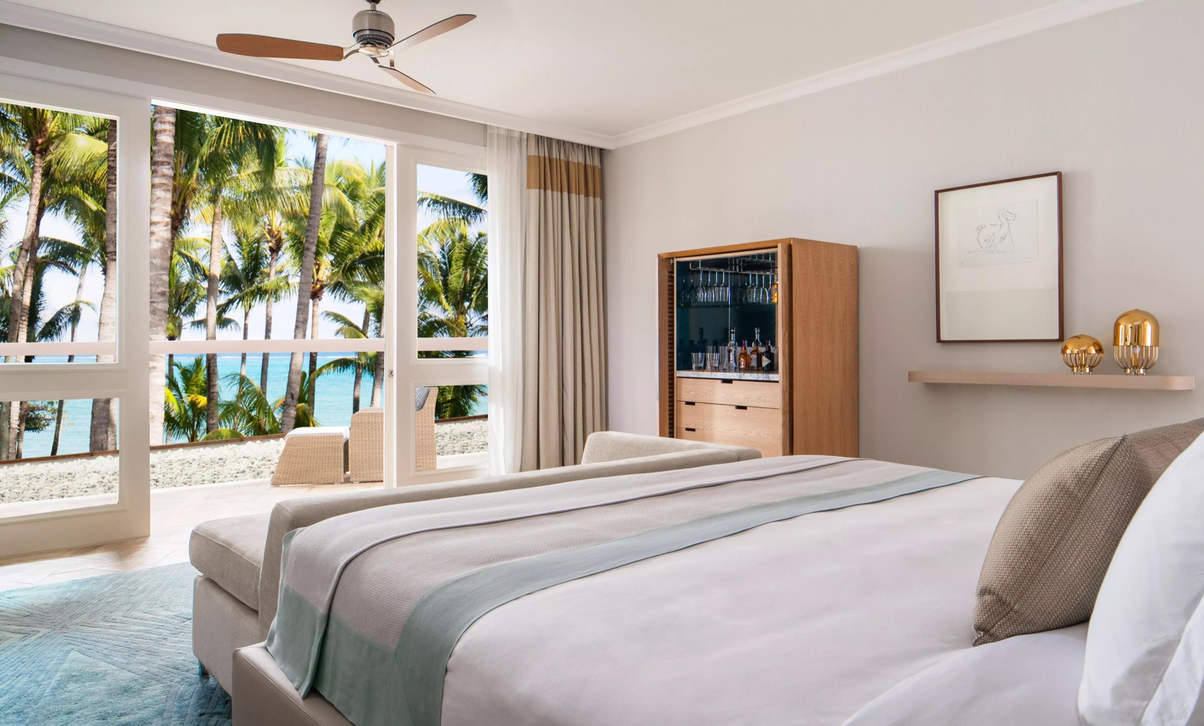 A bedroom with a bed facing glass doors open to a palm-lined terrace and the sea, with a drinks cabinet and a framed print on the wall.