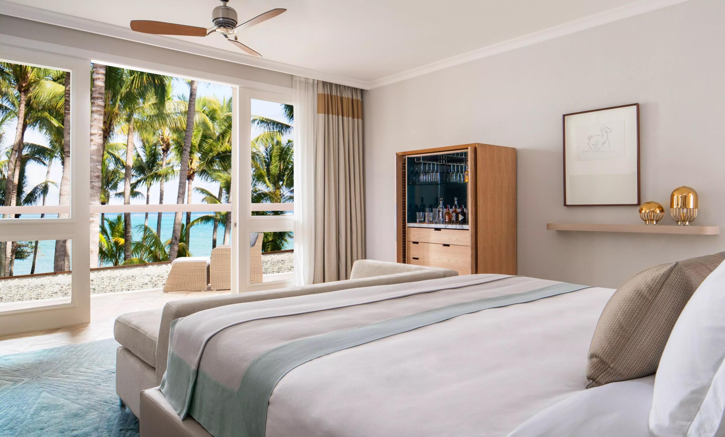 A bedroom with a bed facing glass doors open to a palm-lined terrace and the sea, with a drinks cabinet and a framed print on the wall.