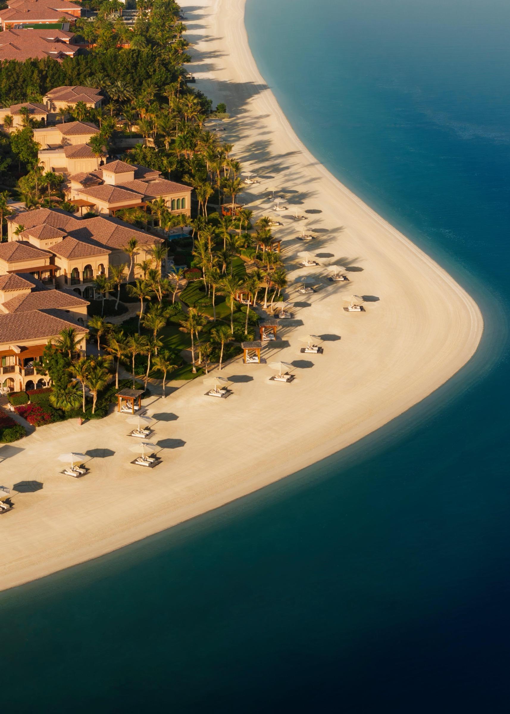 An aerial view of a sandy point lined with palm trees and red-roofed villas, curving into turquoise sea with sunbeds and umbrellas along the shore.