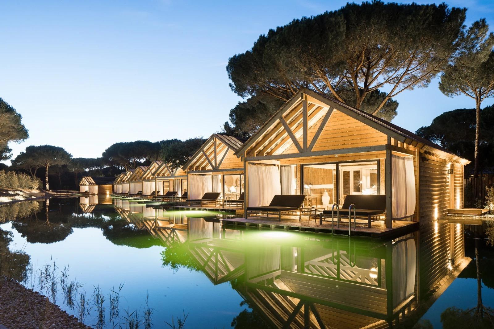 A row of wood cabins on stilts over a still lagoon at dusk, lit from within and reflected in the water, with loungers on their decks.