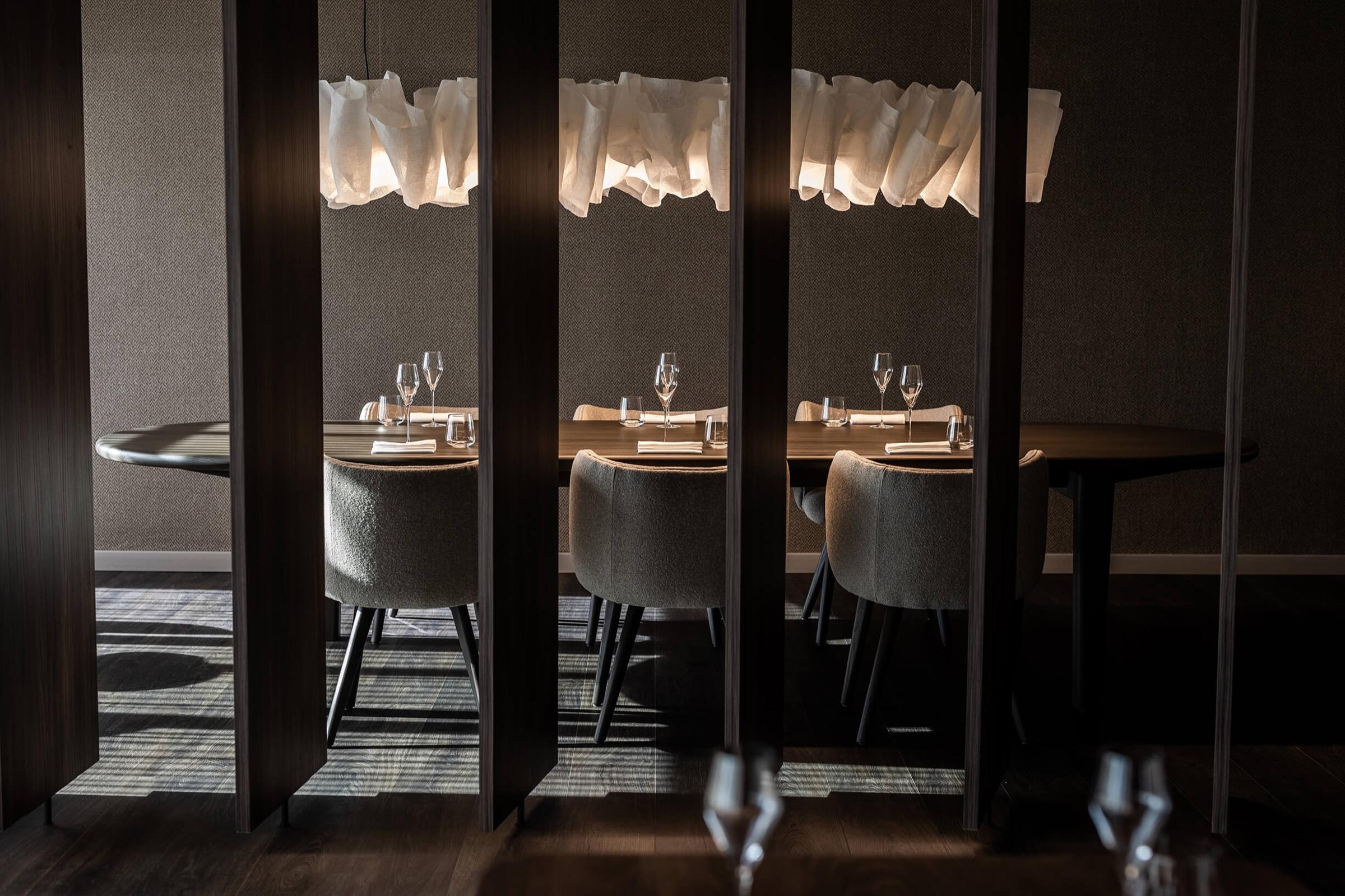 A dark, intimate dining room of tables set with glasses between tall panels, lit by pale hanging lights