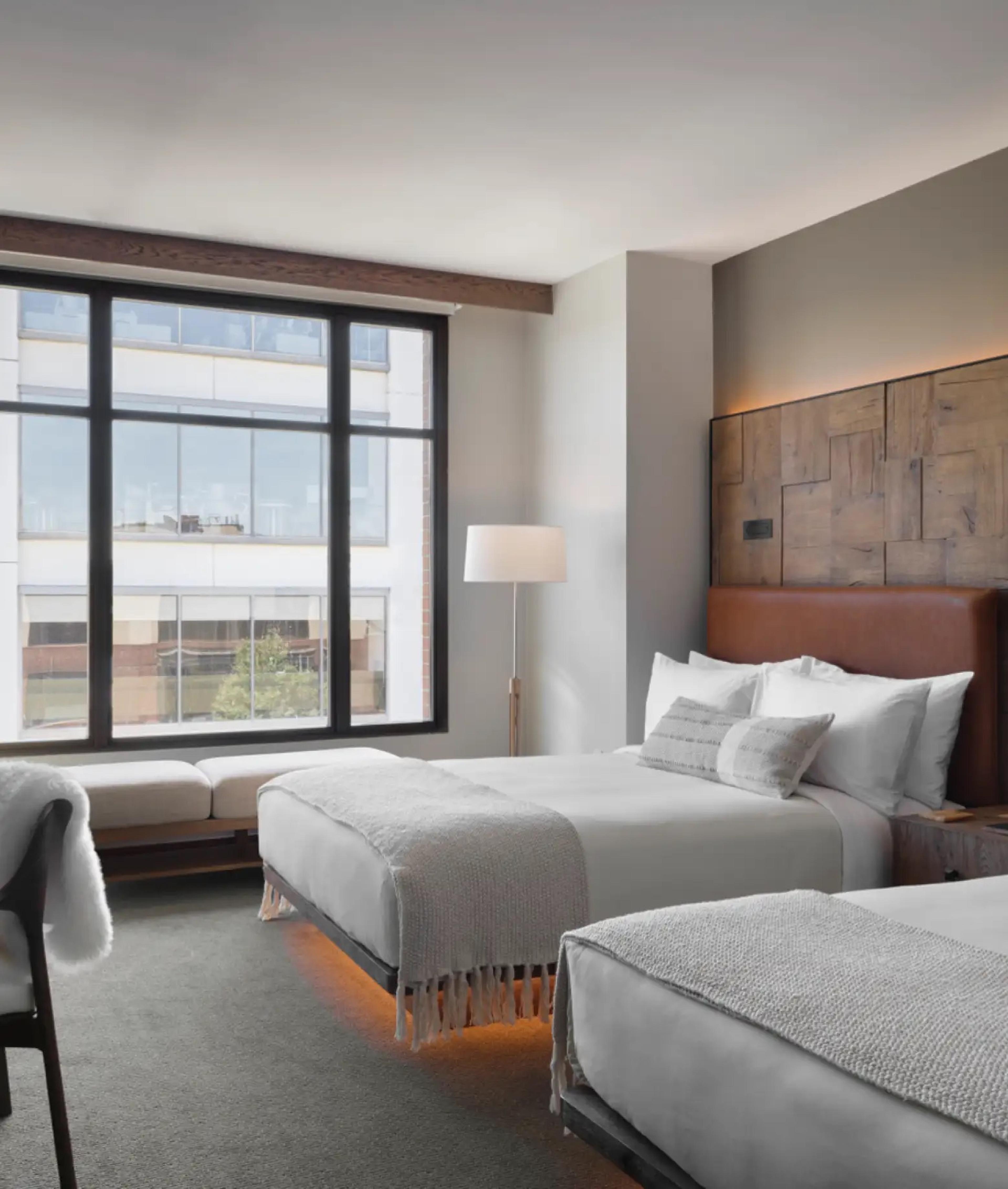 Two queen beds with white bedding and a tan leather headboard, with a bench under a tall window that looks onto a pale office building.