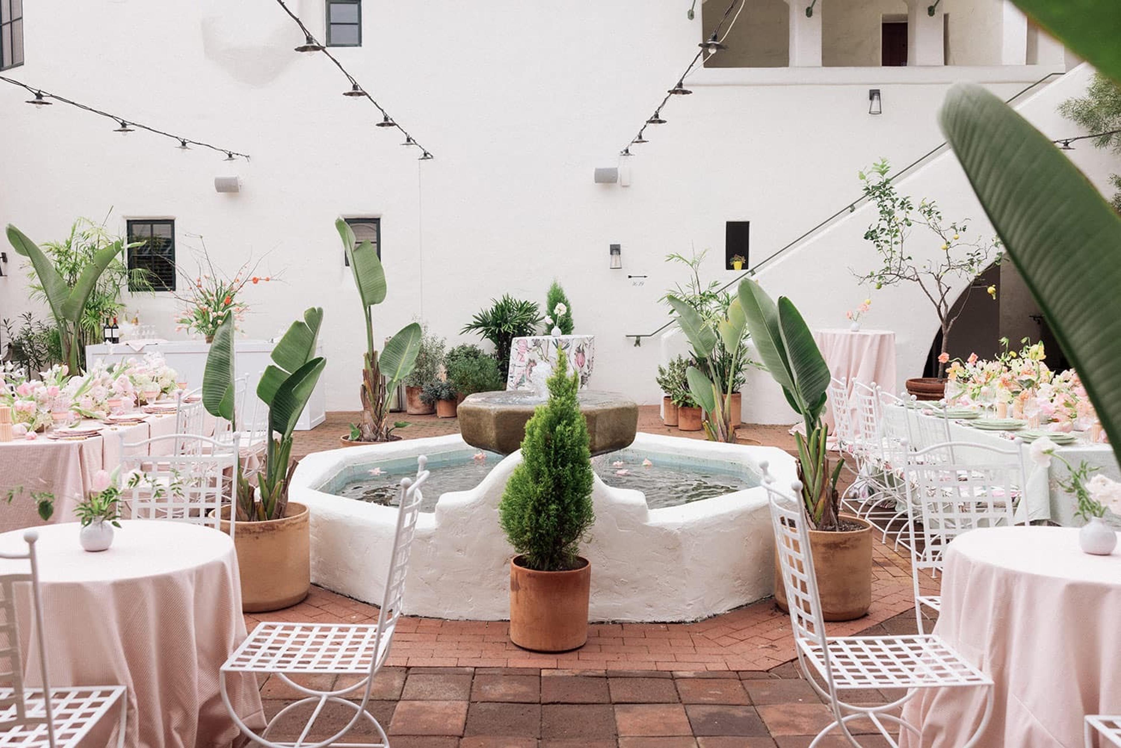 An outdoor courtyard with round tables in pink linens and white folding chairs set around a stone fountain, strung with lights beneath a white building's balcony.