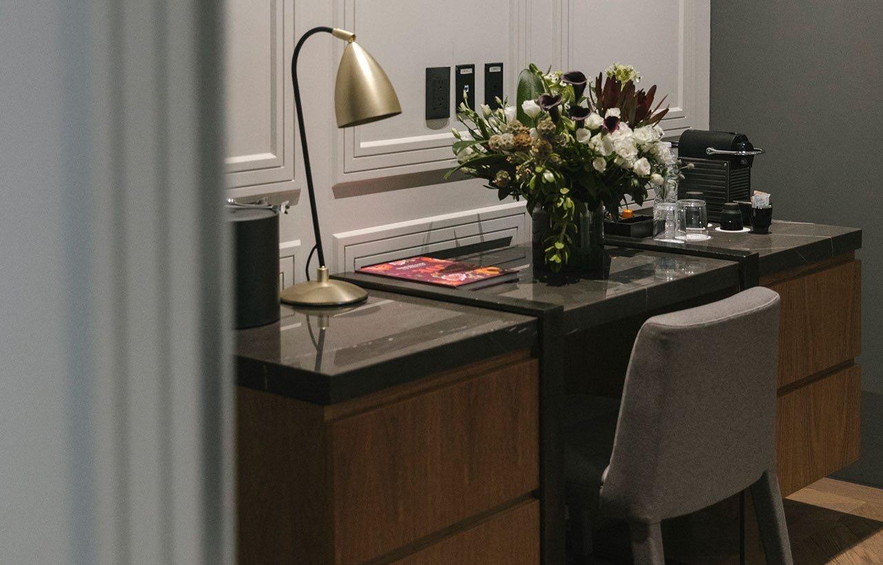 A hotel room desk with a brass lamp, a vase of white flowers, a coffee machine and glassware, beside an upholstered chair.
