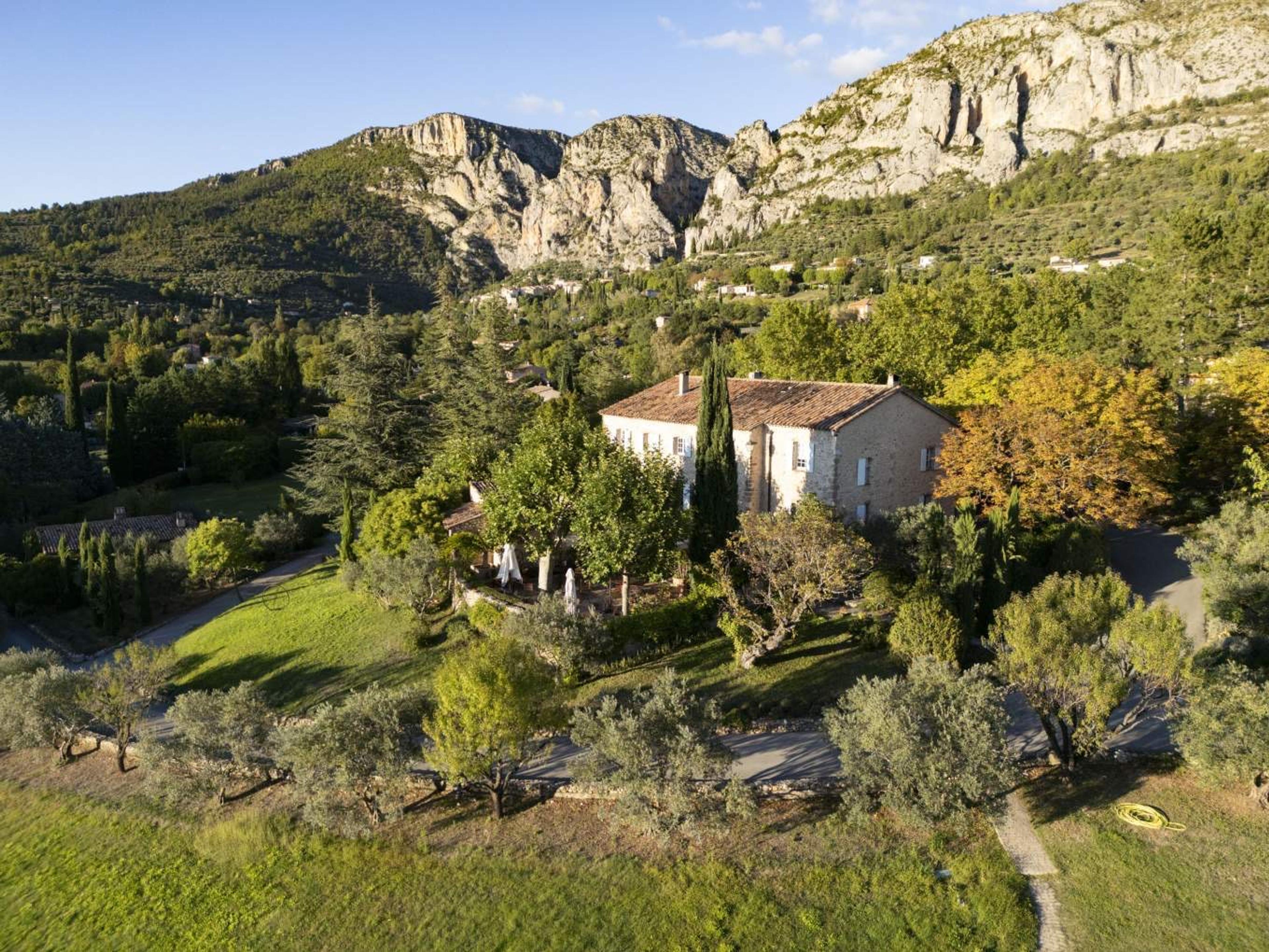 An aerial view of a stone farmhouse and olive groves set below steep limestone cliffs, lit by low evening sun.