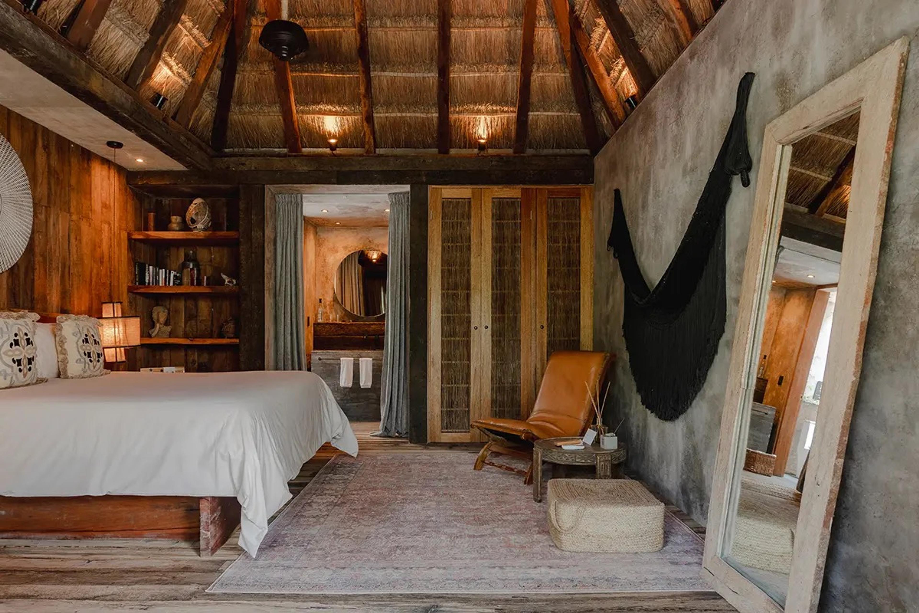 A bedroom with a palapa thatched ceiling, a white bed, a leather chair and a tall freestanding mirror.