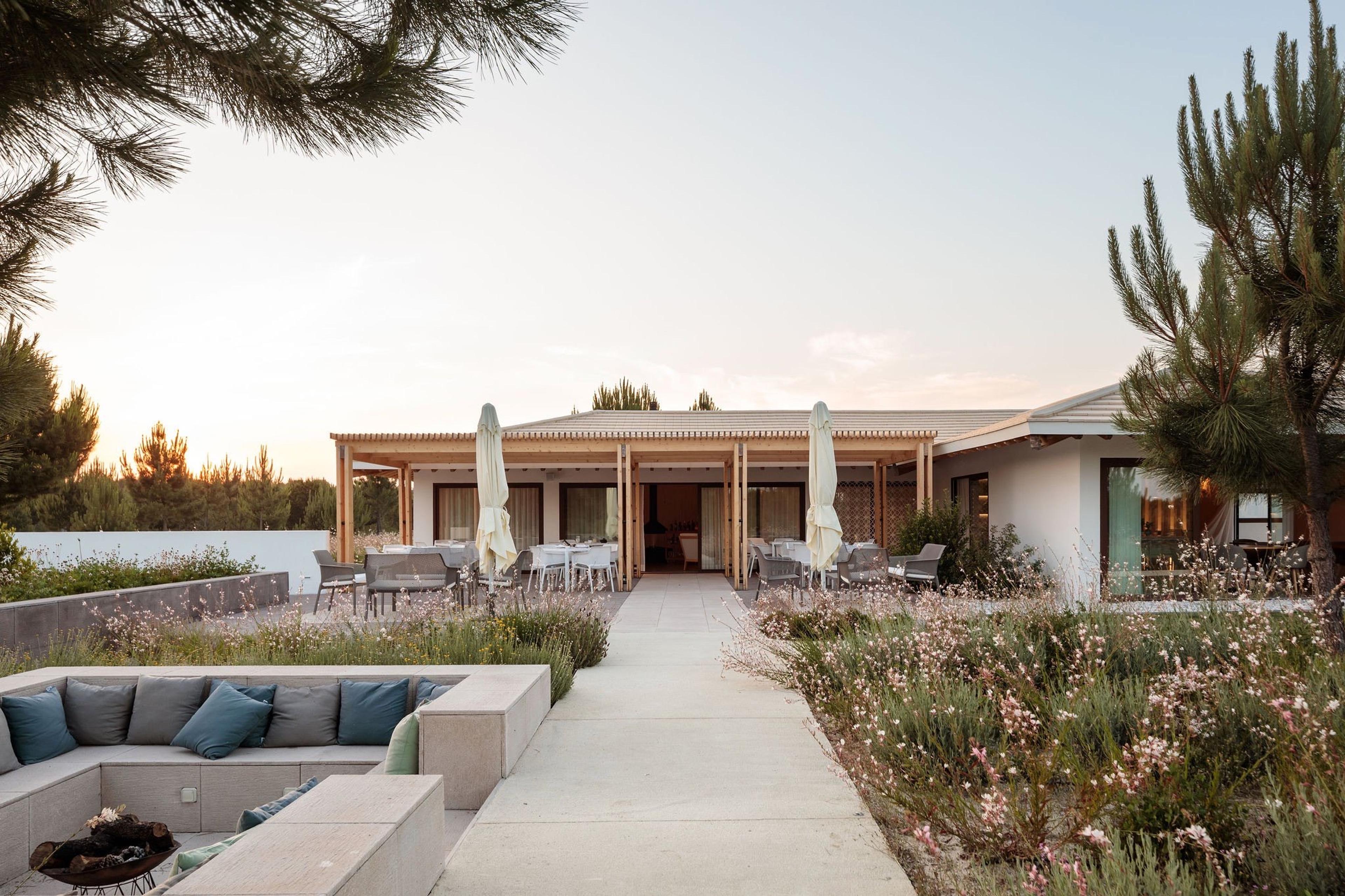 A pergola-covered terrace with dining tables and a built-in cushioned seating area around a firepit, flowering grasses in the foreground at dusk.