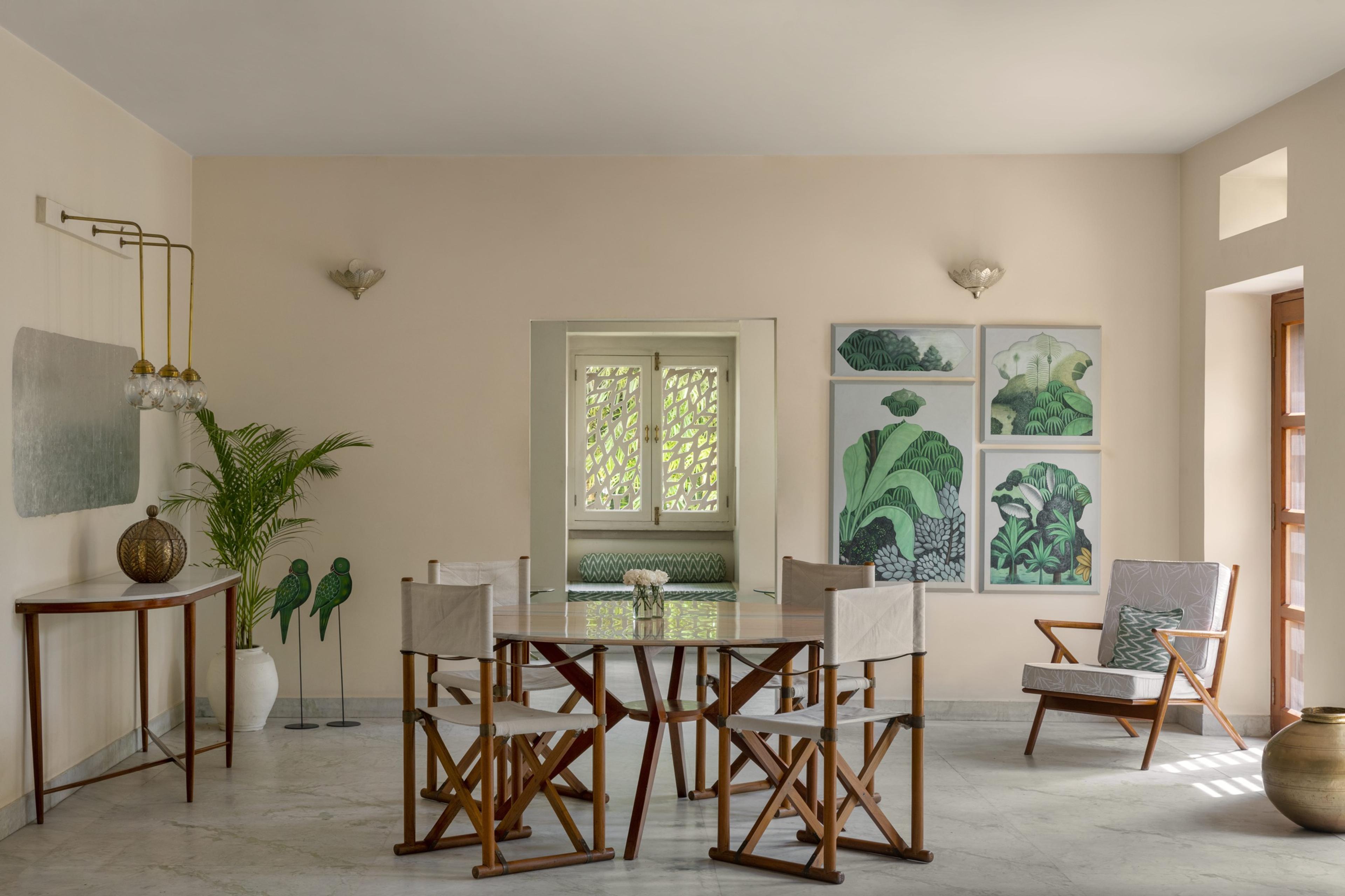 A round dining table with director's chairs set in a plaster-walled room, with potted palms, botanical prints and a console table against the wall.